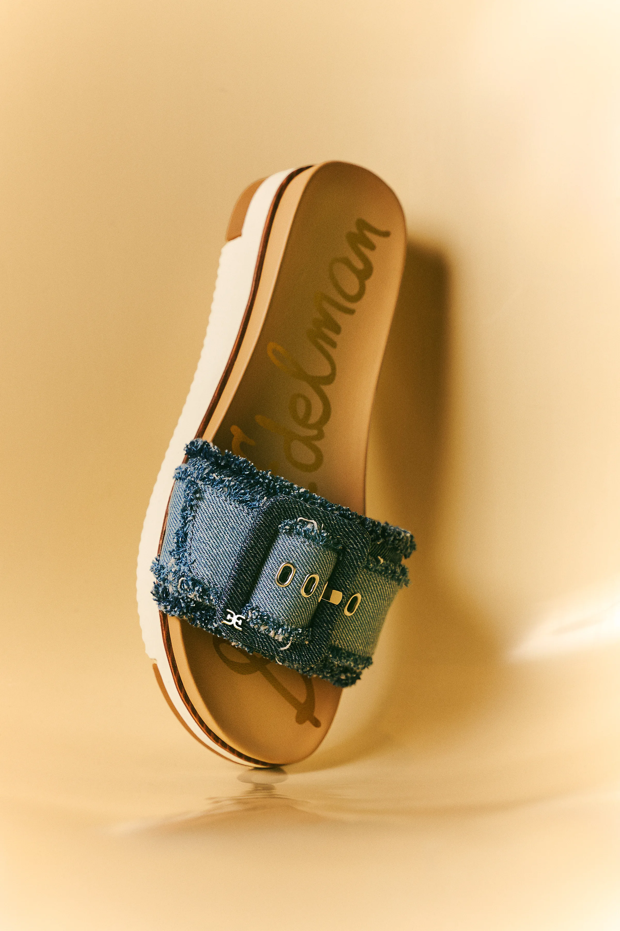 Frayed denim buckle slide sandal with white platform sole on a warm golden reflective backdrop, for Sam Edelman.