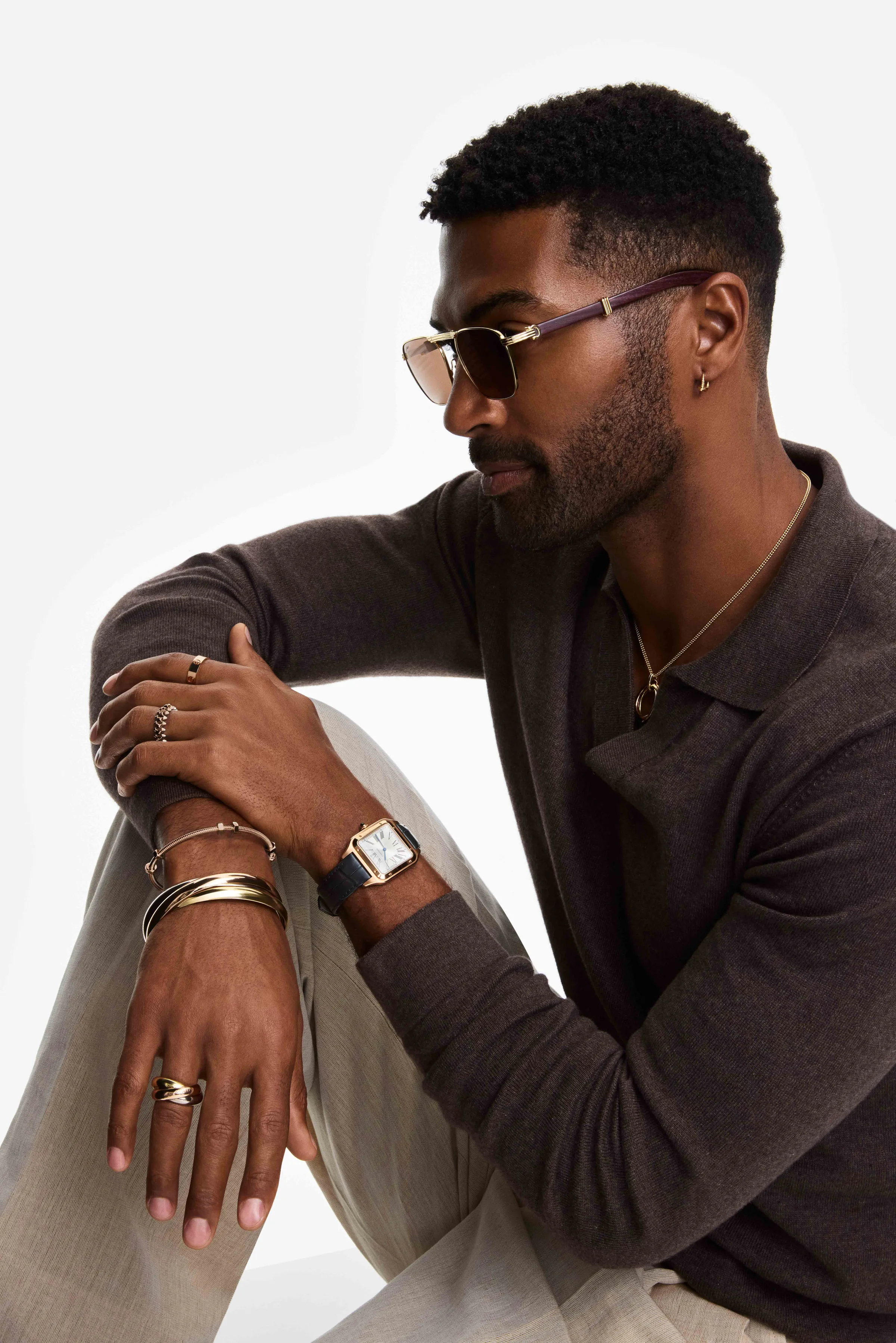 Seated man in brown knit polo and sunglasses wearing a gold watch, stacked bracelets and rings on white, for Cartier.