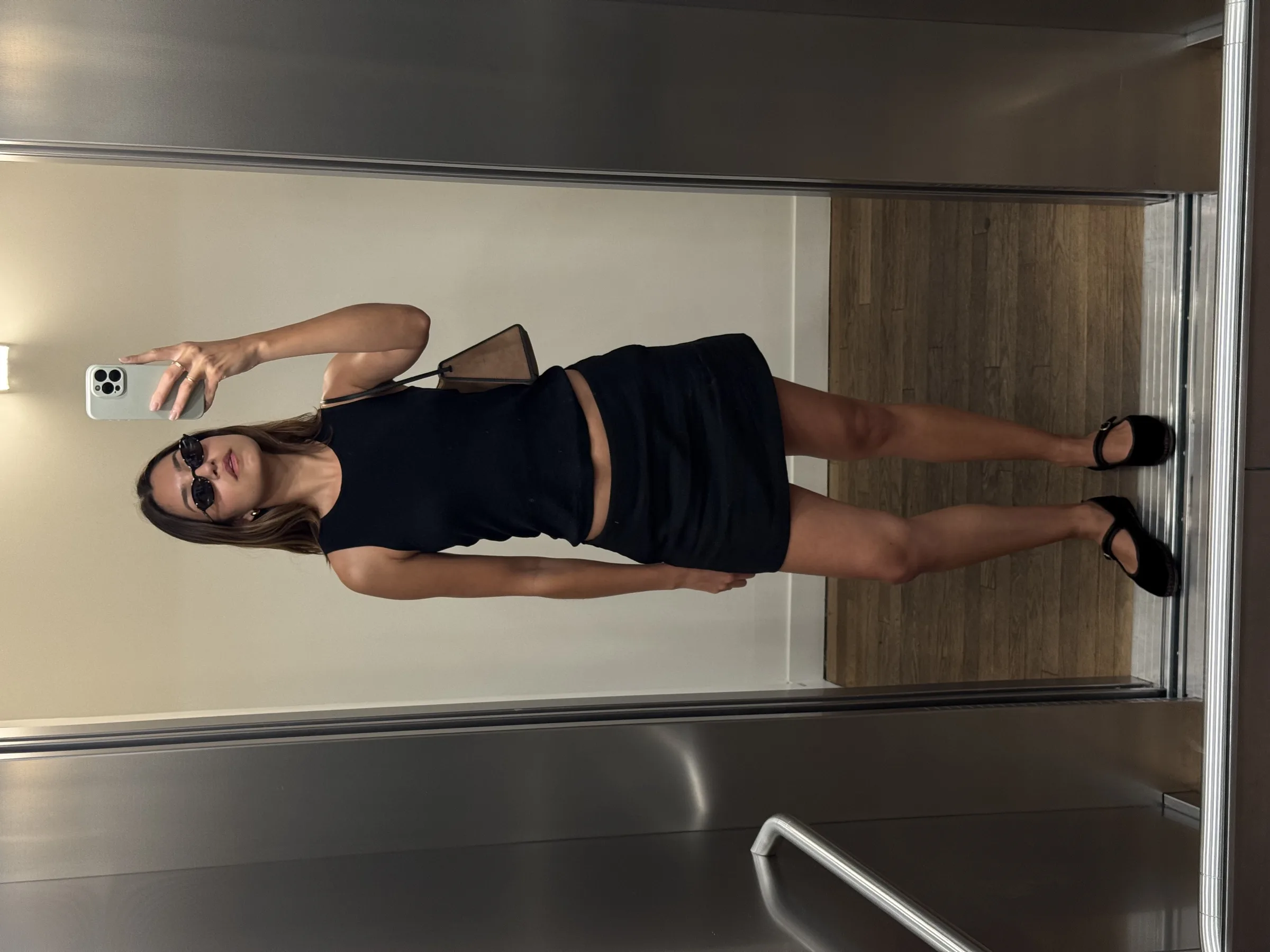 Woman taking a mirror selfie in a steel elevator, wearing a black tank, mini skirt, sunglasses and Mary Jane flats.