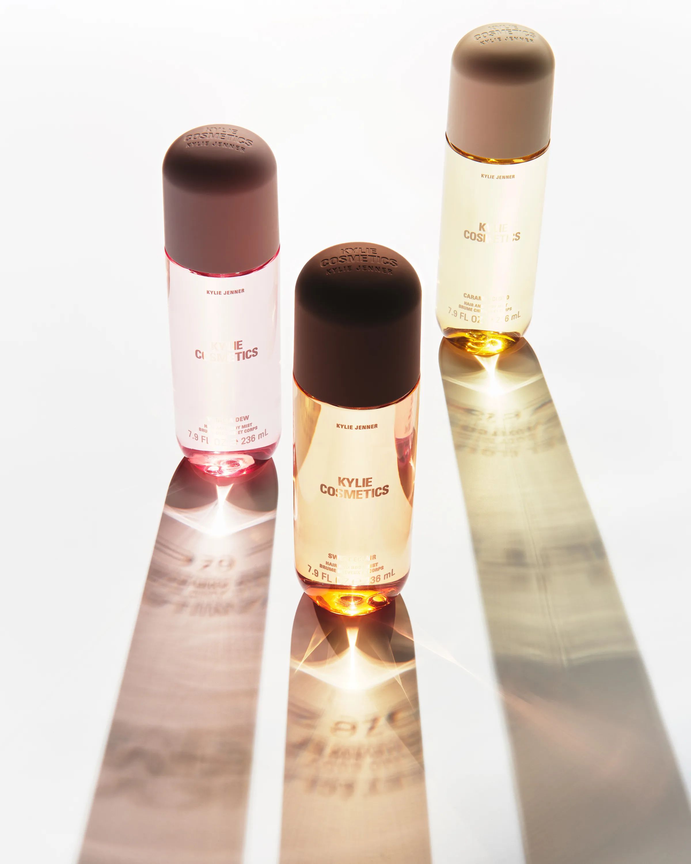 Three hair and body mist bottles with brown caps casting long sunlit shadows on a white surface, for Kylie Cosmetics.