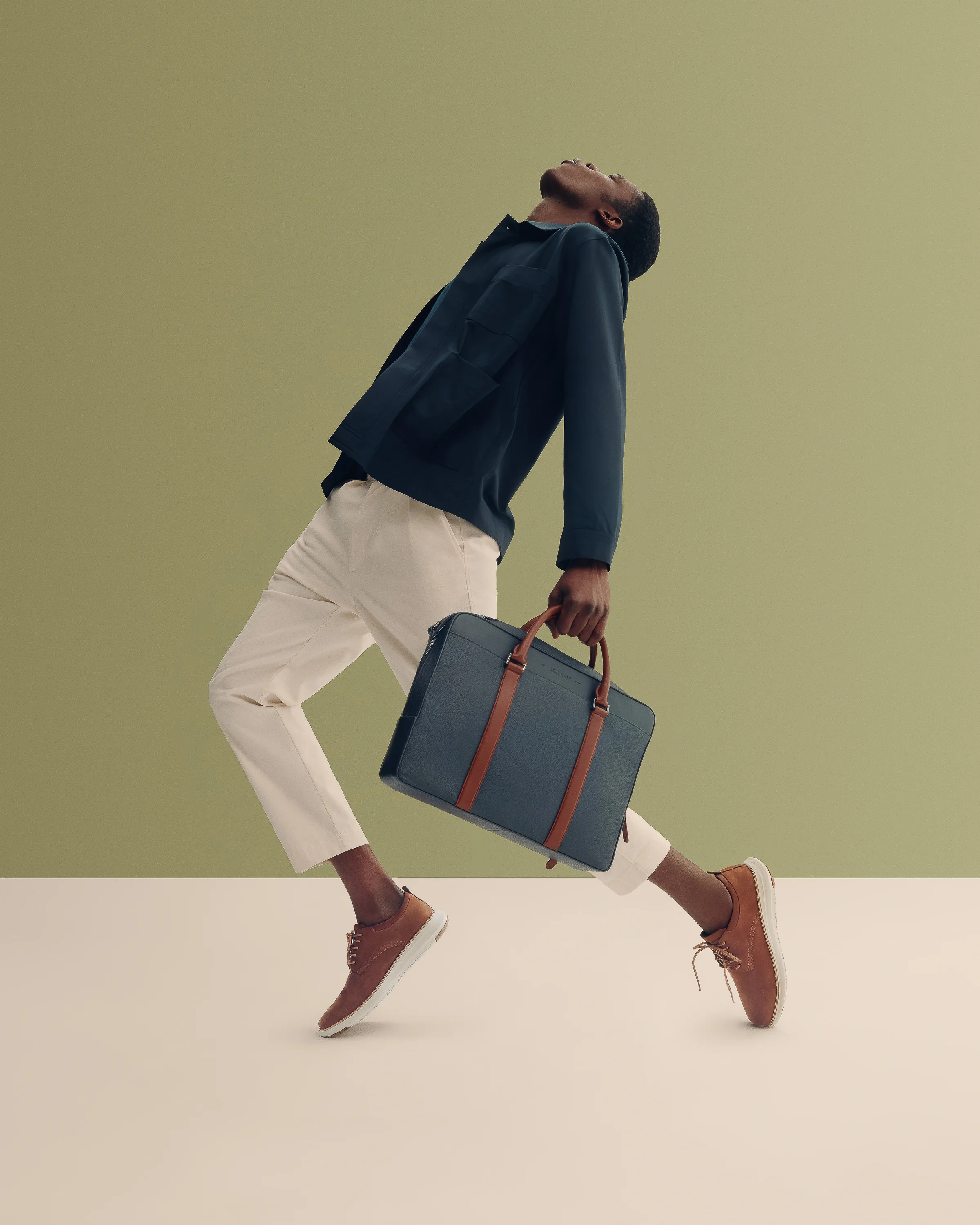 Man mid-stride with head tilted back, carrying a navy briefcase in tan sneakers against an olive backdrop, for Cole Haan.