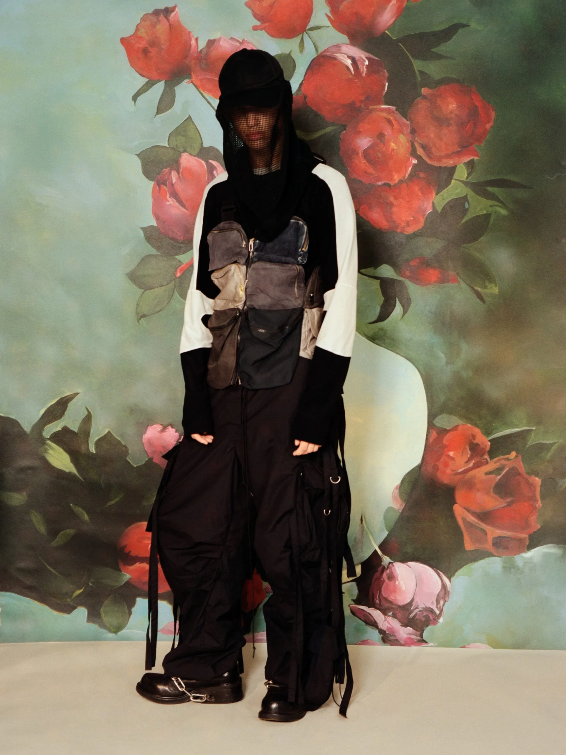 Model in black hood, patchwork utility vest and strapped cargo pants before a painted rose backdrop, for Office Magazine.