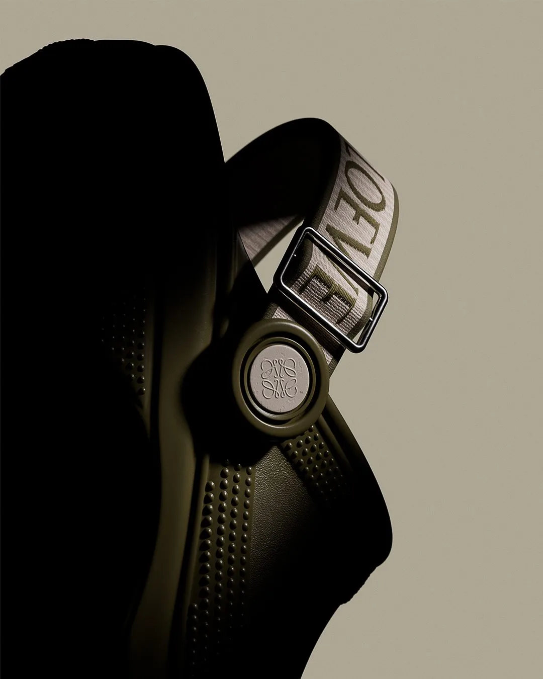 CGI render of an olive green clog with a logo strap, metal buckle and anagram button on a beige background, for Loewe.