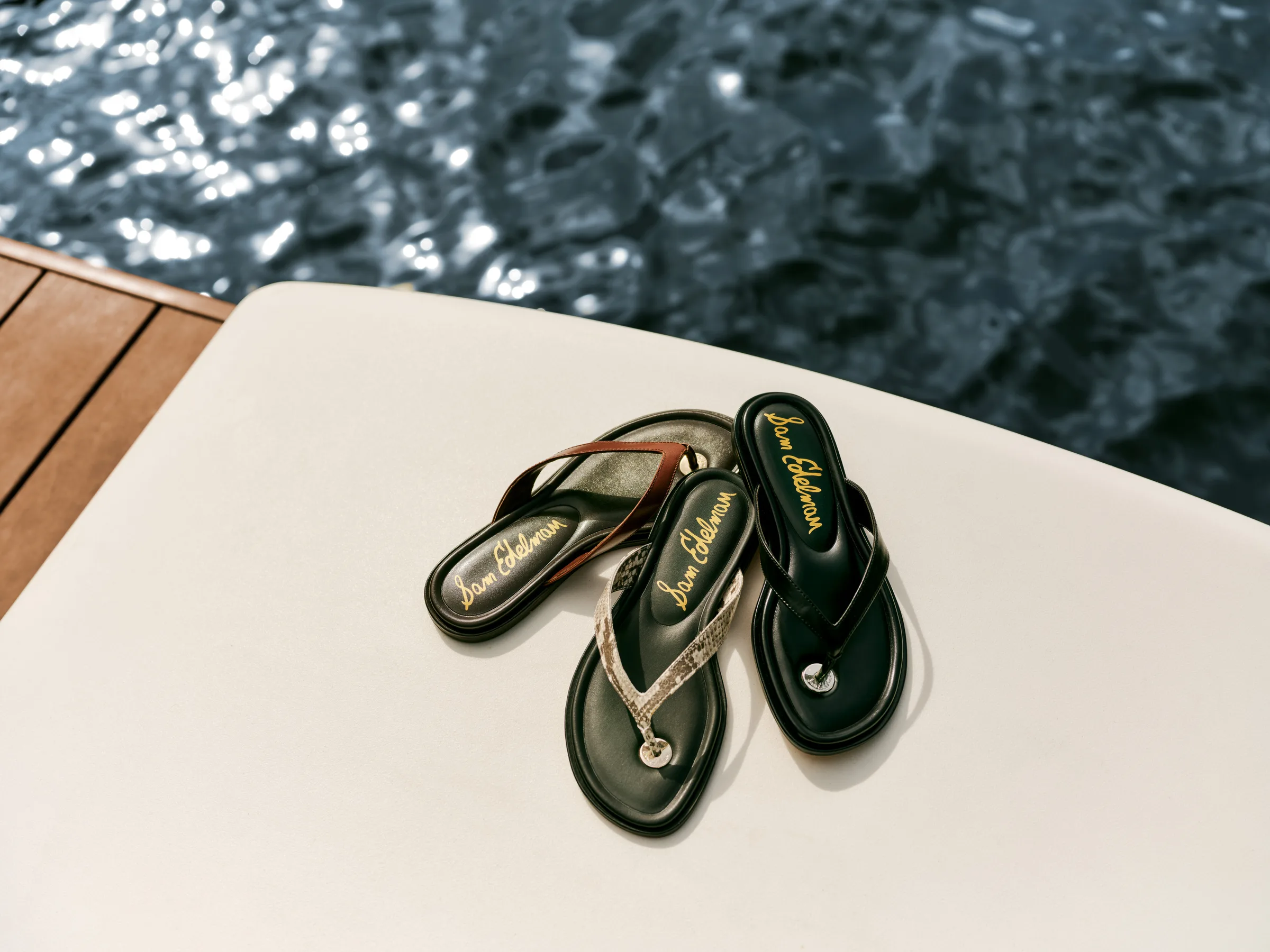 Three thong sandals in brown, snakeskin and black on a white boat deck above sparkling water, for Sam Edelman.