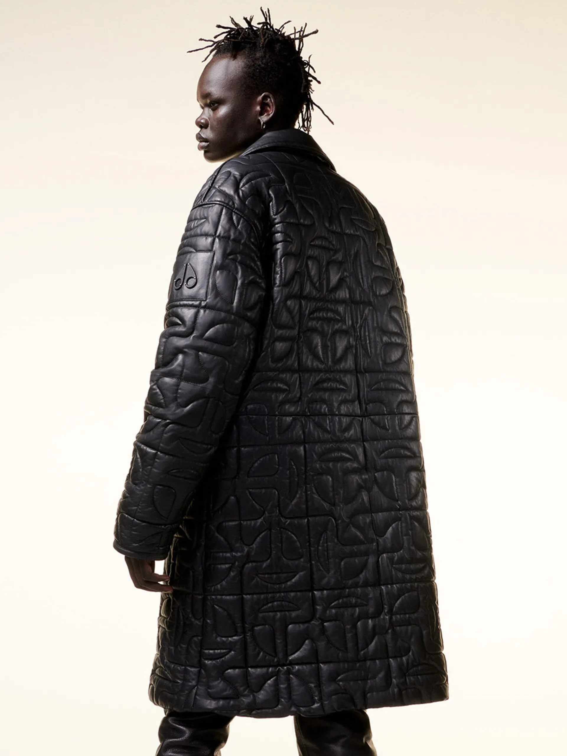 Model seen from behind in a long black leather coat quilted with a logo pattern, on a cream backdrop, for Moose Knuckles.