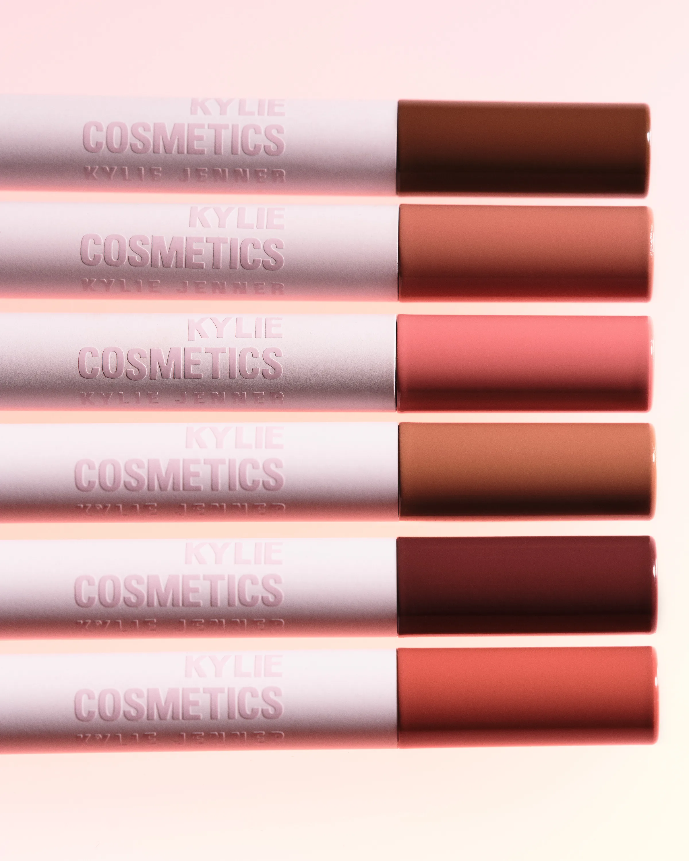 Six lip liners with pink barrels and nude, pink and brown tips stacked in a row on a pale pink background, for Kylie Cosmetics.