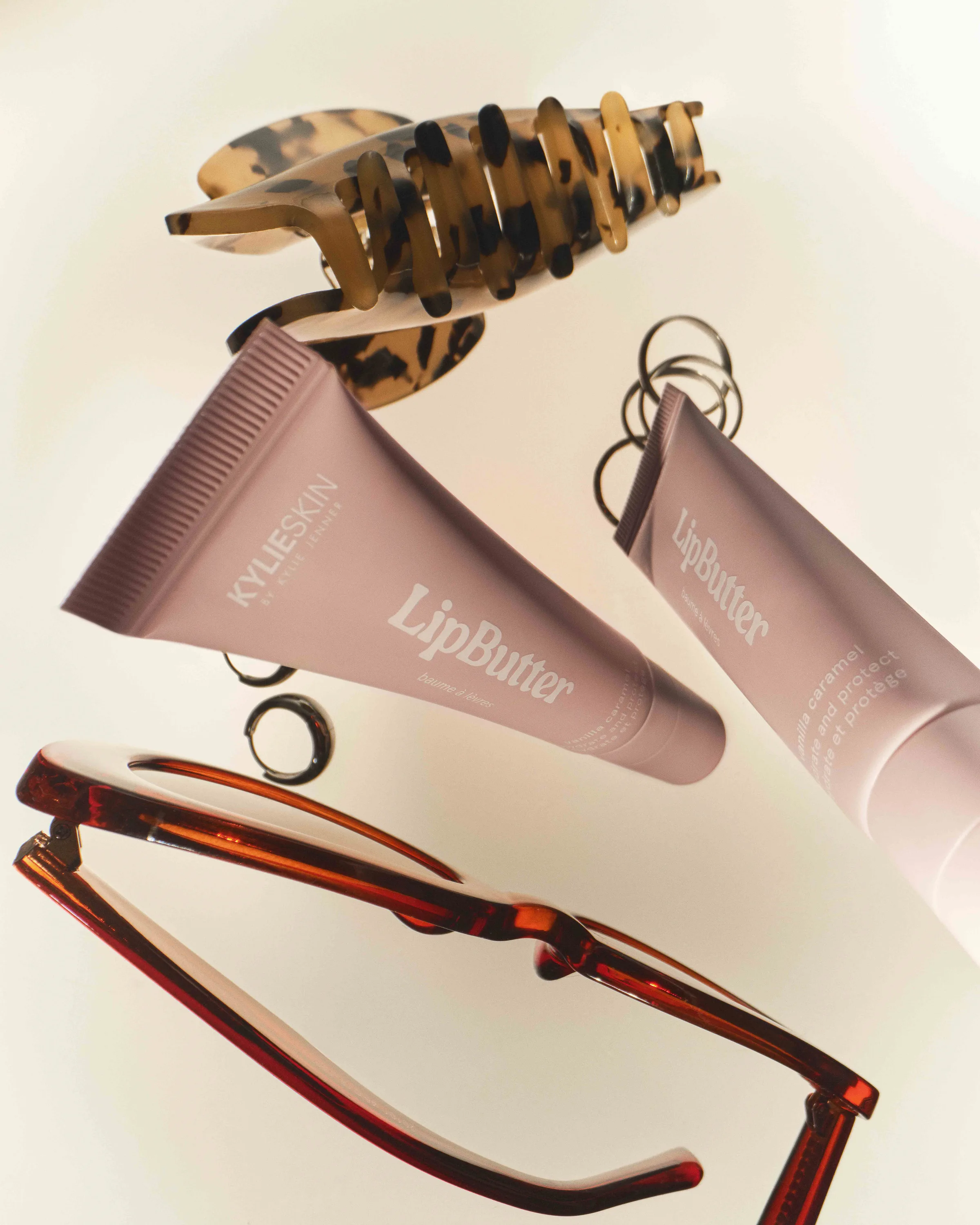 Mauve Kylie Skin Lip Butter tubes floating with a tortoiseshell claw clip, rings and red sunglasses, for Kylie Cosmetics.