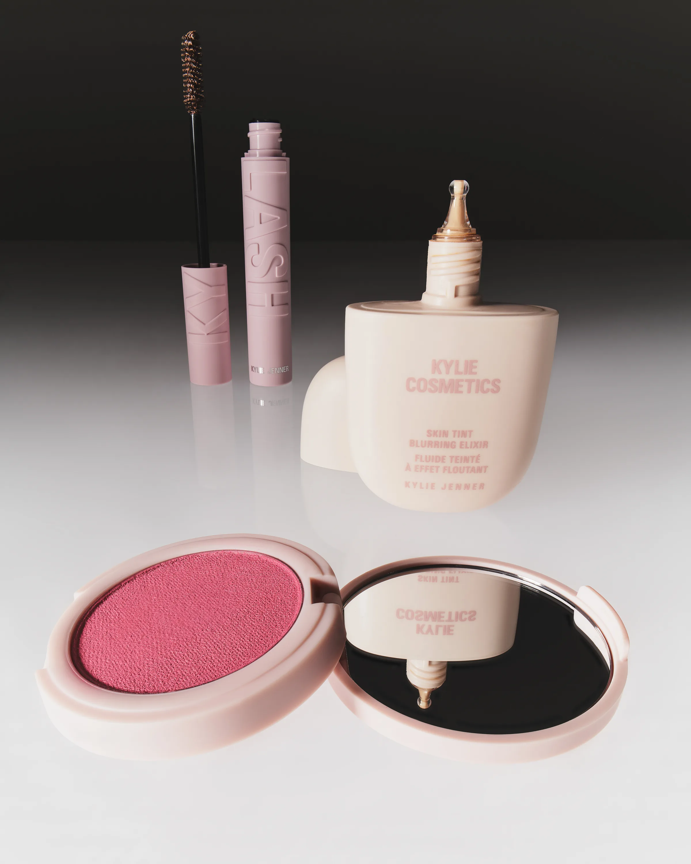 Skin tint bottle, pink Kylash mascara and open blush compact with mirror on a glossy white surface, for Kylie Cosmetics.