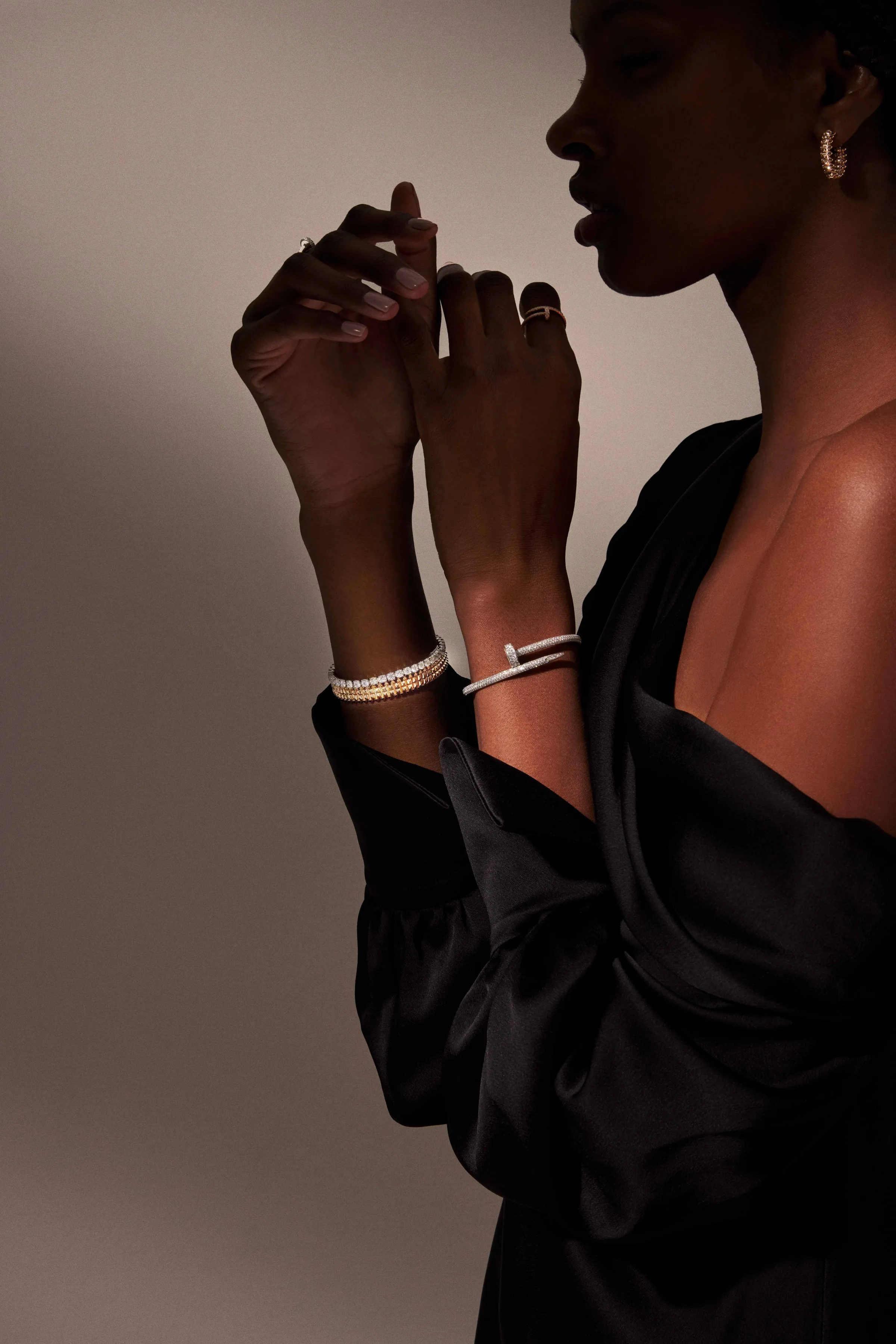 Model in black satin dress wearing diamond bracelets, a nail bangle, rings and hoop earrings in soft side light, for Cartier.