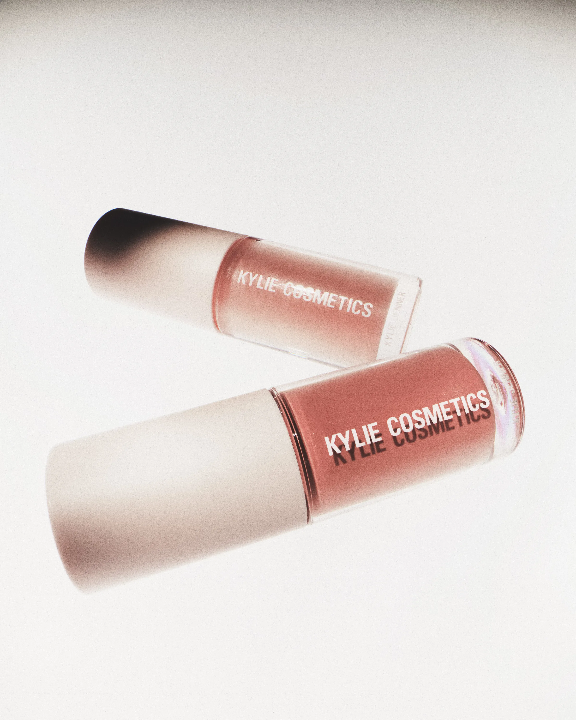 Two pink lip gloss tubes with white caps angled on a white reflective surface, for Kylie Cosmetics.