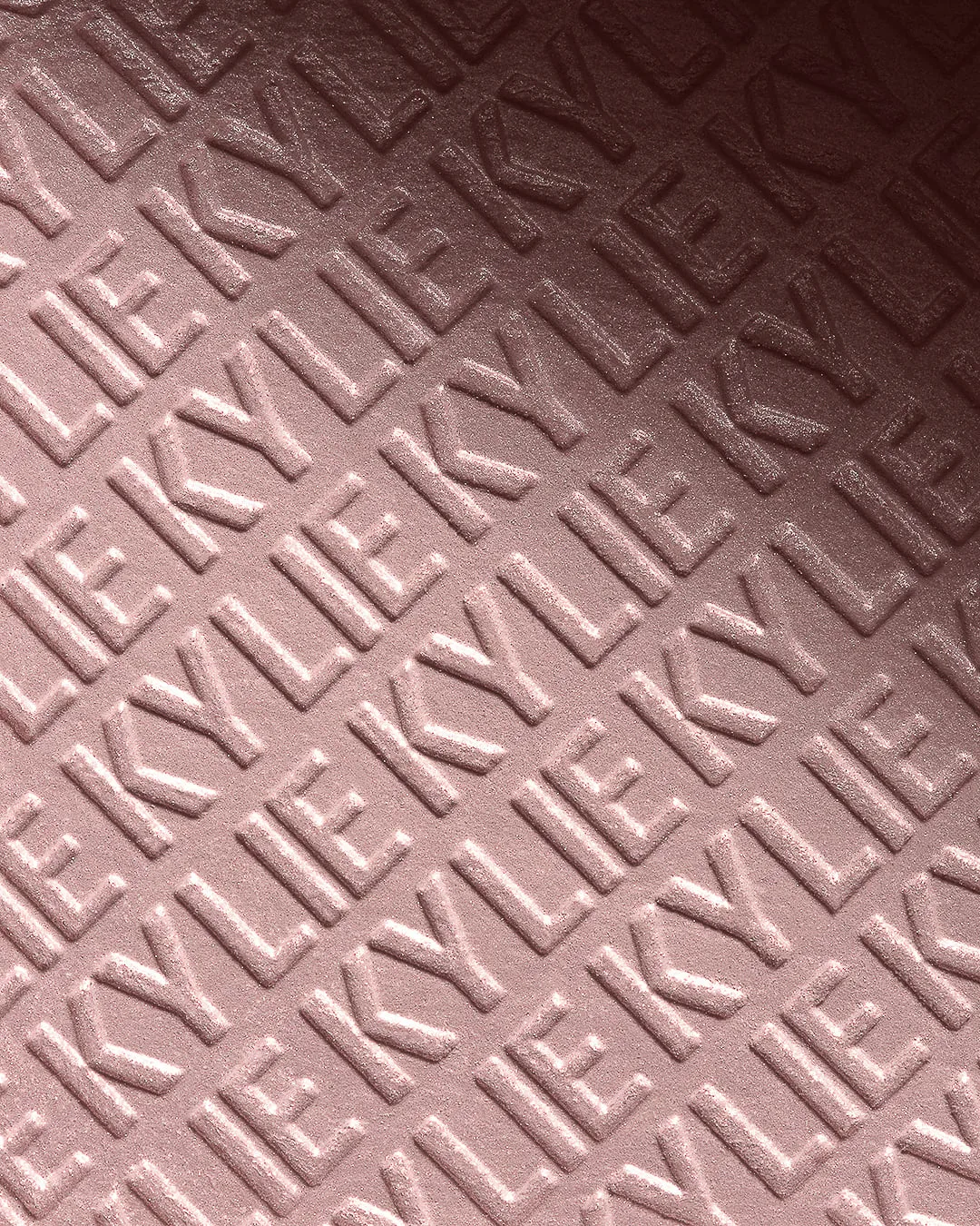 Close-up of pink pressed powder embossed with a repeating KYLIE pattern under raking light, for Kylie Cosmetics.