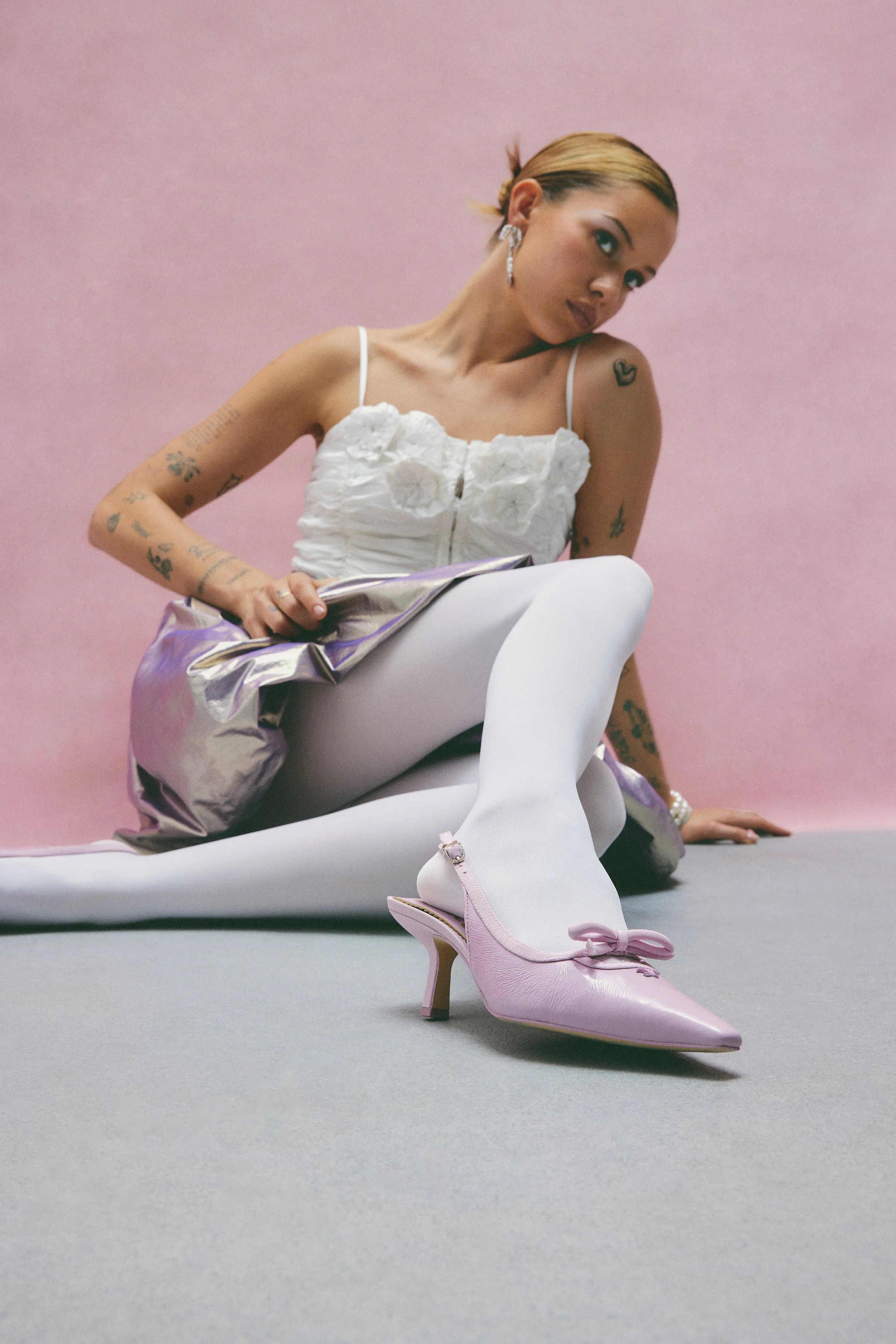 Model in white tights and lace top seated on grey floor, wearing pale orchid bow slingback heels, for Sam Edelman.