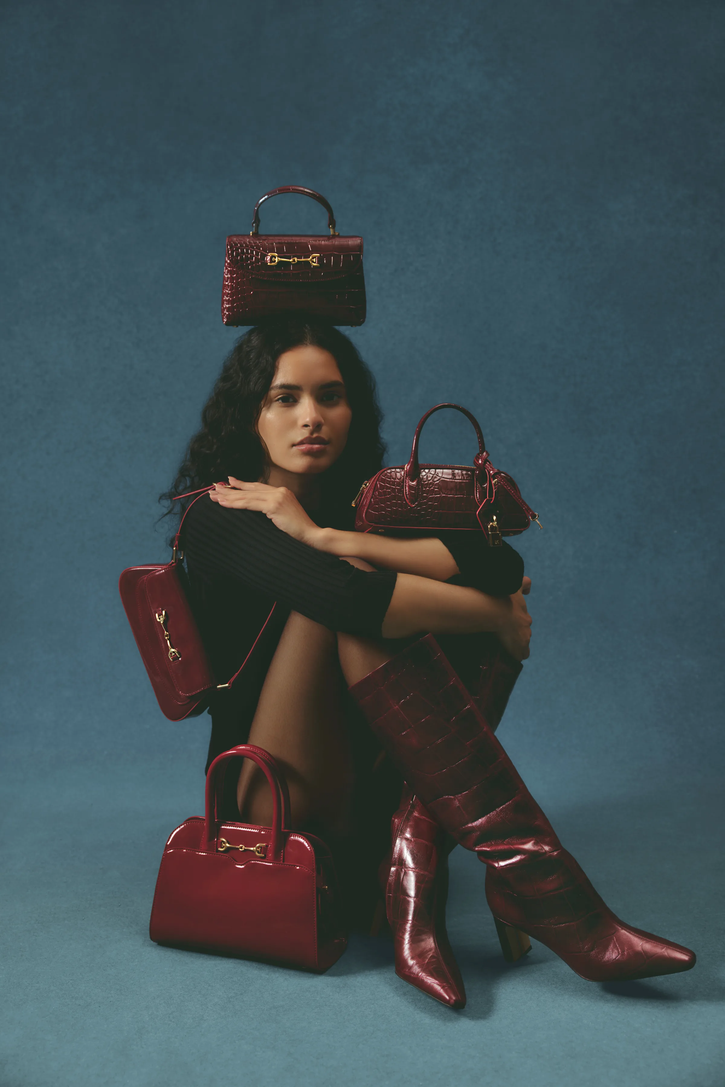 Model seated on teal backdrop with merlot croc-embossed handbags and knee-high boots, one bag on her head, for Sam Edelman.
