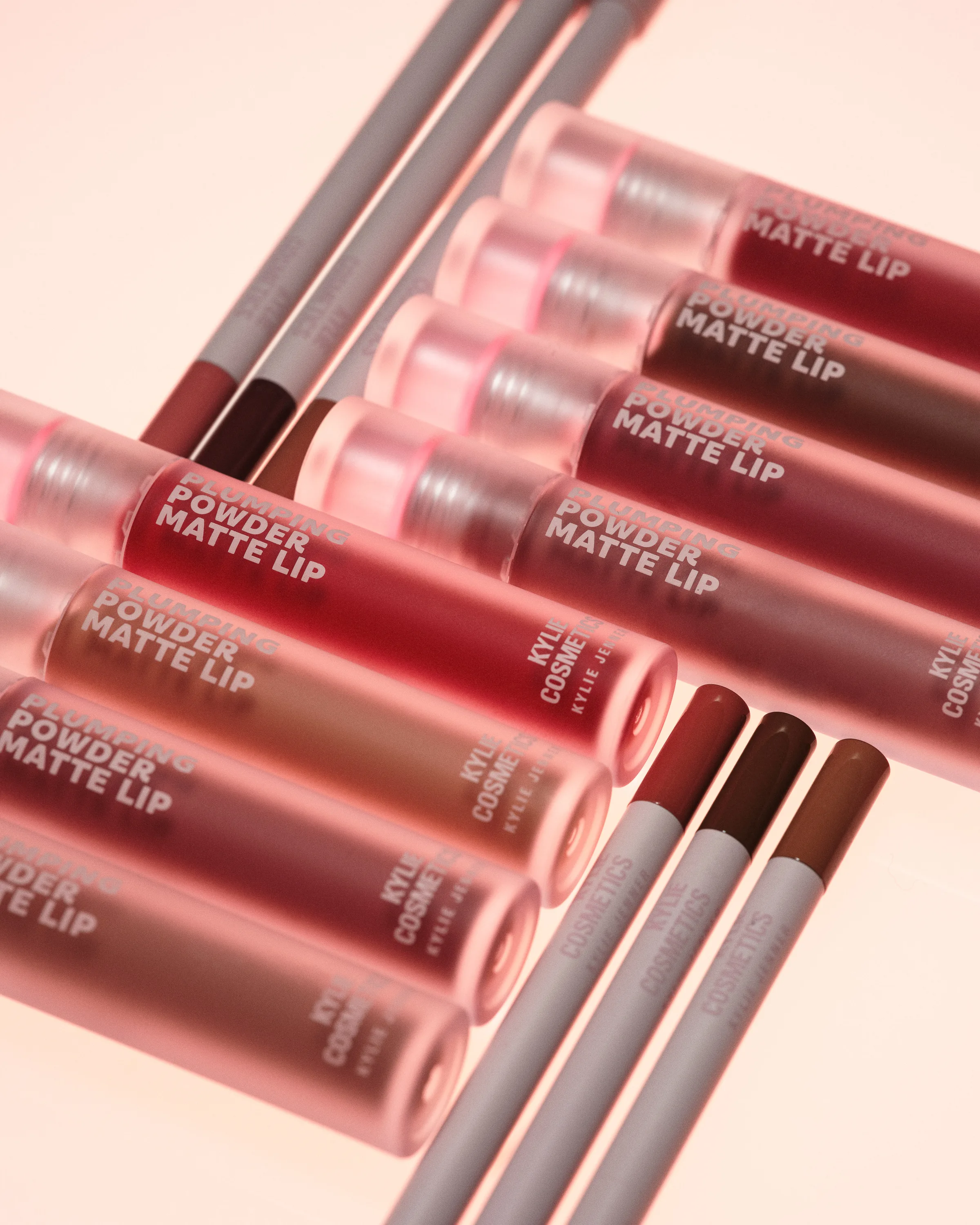 Rows of pink frosted Plumping Powder Matte Lip tubes and lip liners in nude and berry shades on a peach backdrop, for Kylie Cosmetics.