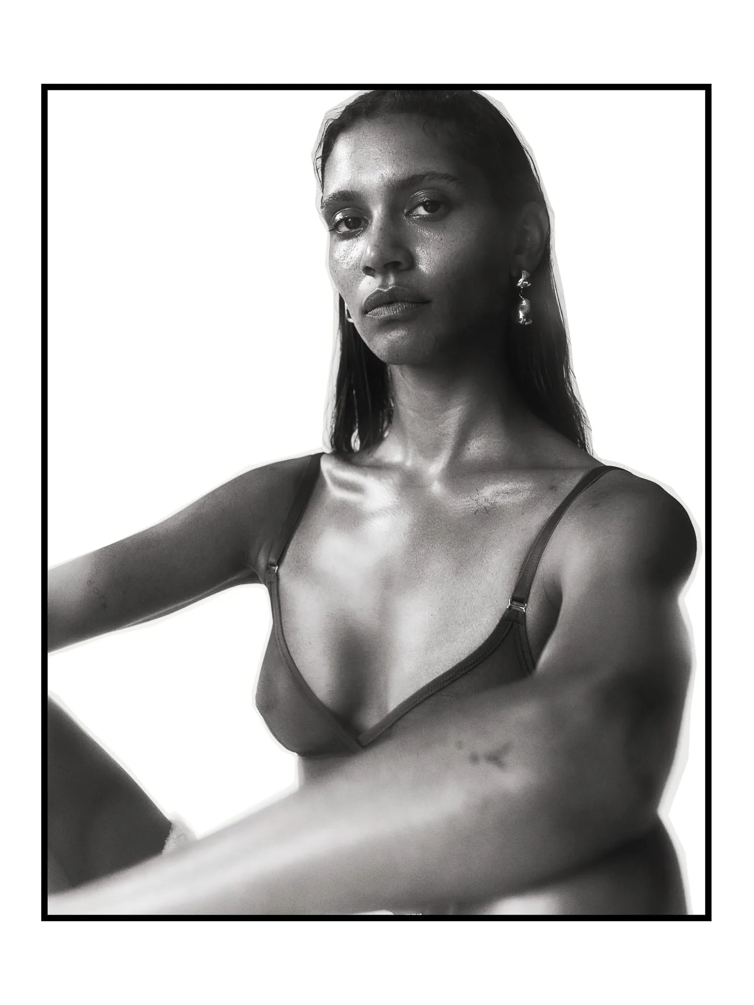 Black and white portrait of a model in a thin-strap bralette and drop earrings, seated against white, for The Last Magazine.