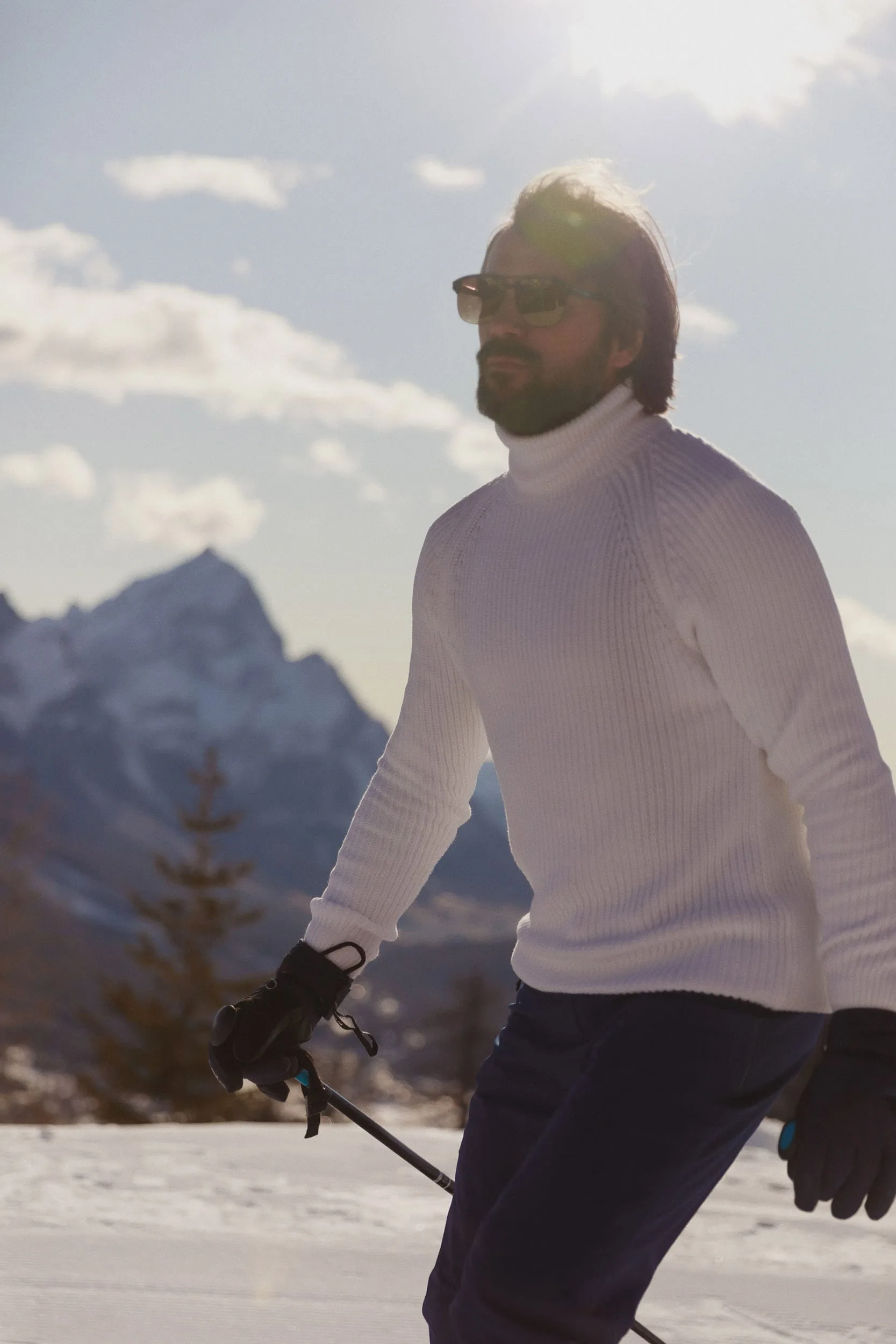 Bearded man in sunglasses and white ribbed turtleneck skiing with poles before snowy mountain peaks, for Marriott.