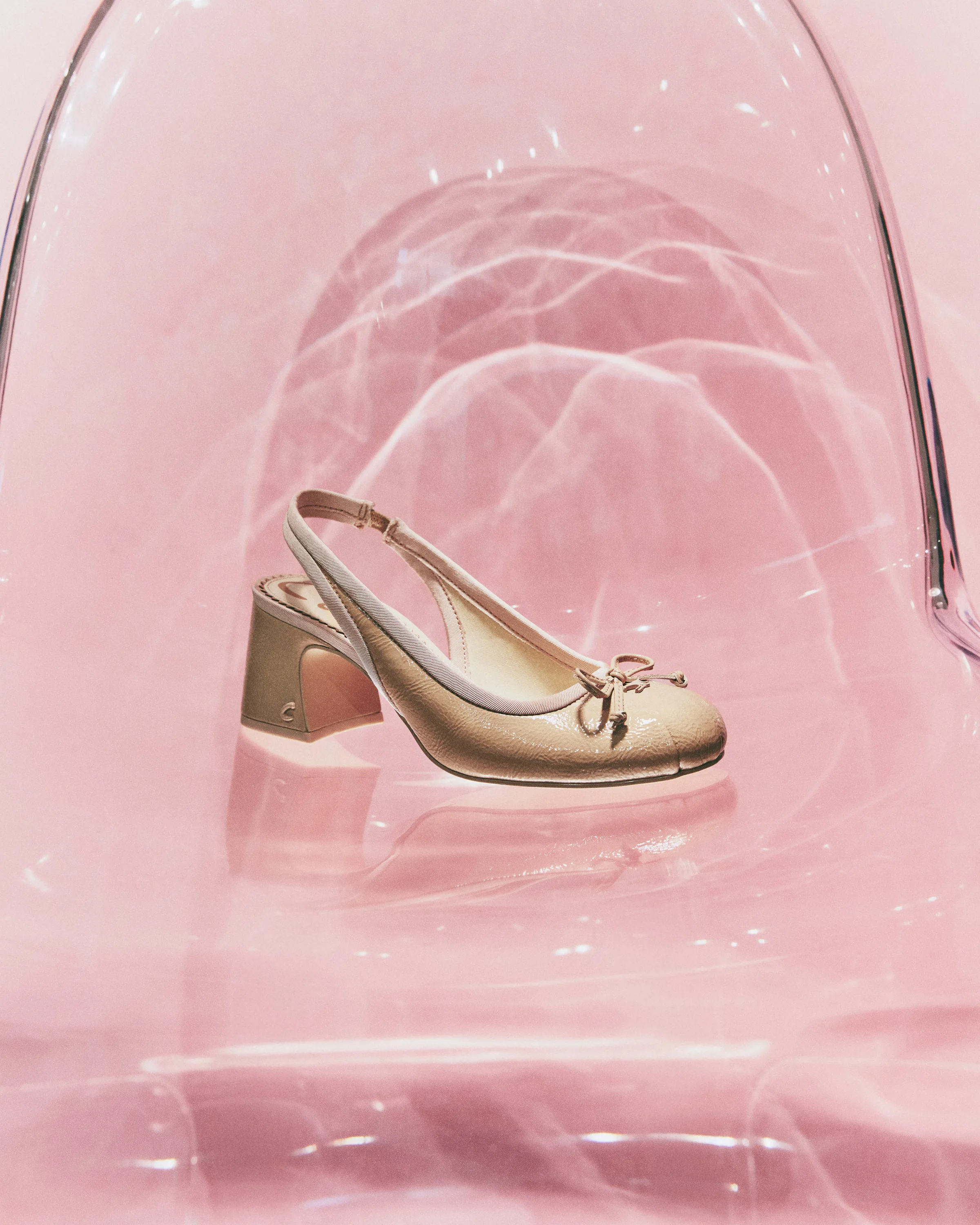 Vanilla patent slingback heel with a bow toe, set inside a clear acrylic chair on a pink backdrop, for Sam Edelman.