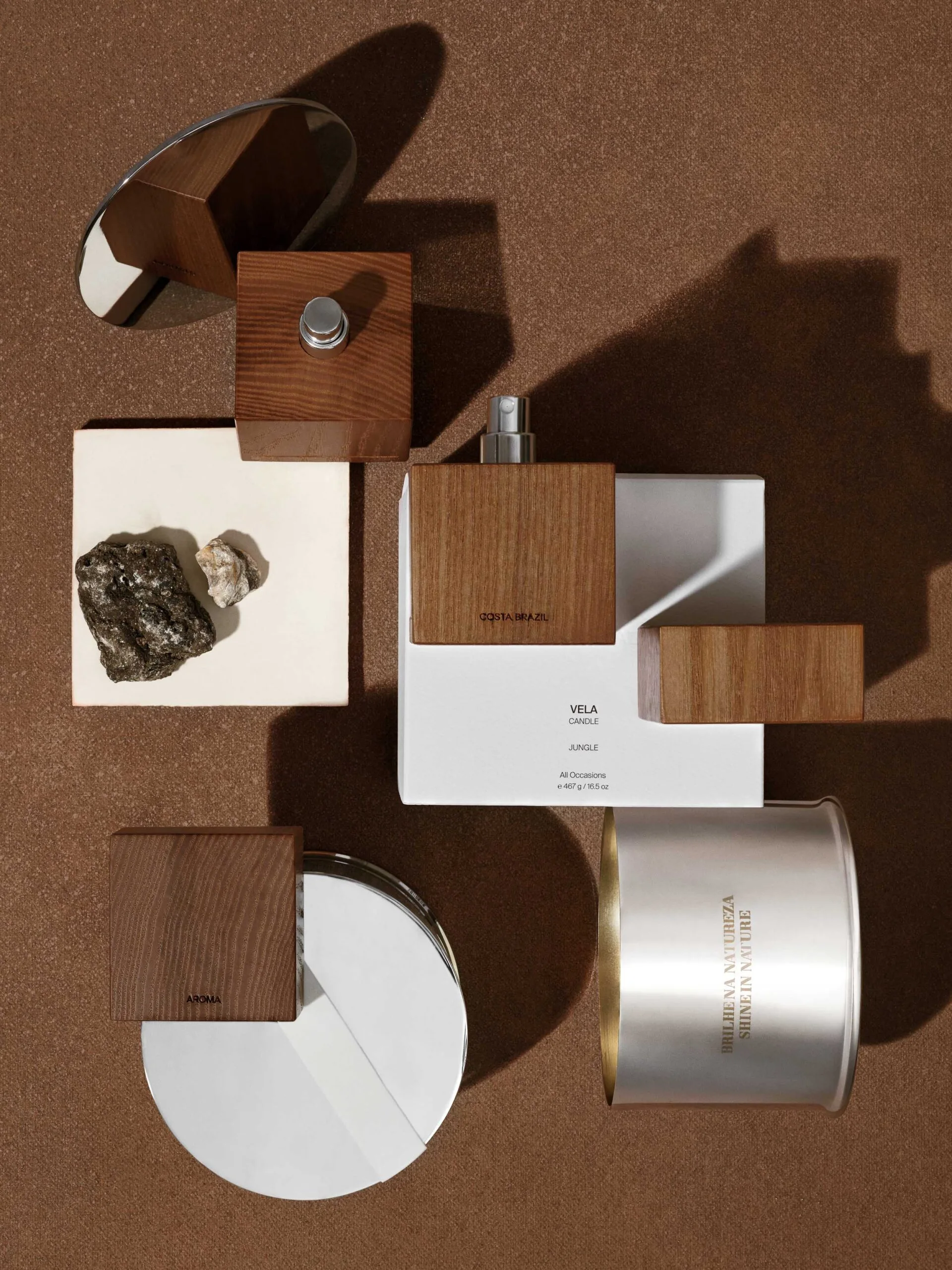 Flatlay of wood-capped perfume, Vela candle box, silver candle tins and stones on brown felt in hard sunlight, for Costa Brazil.