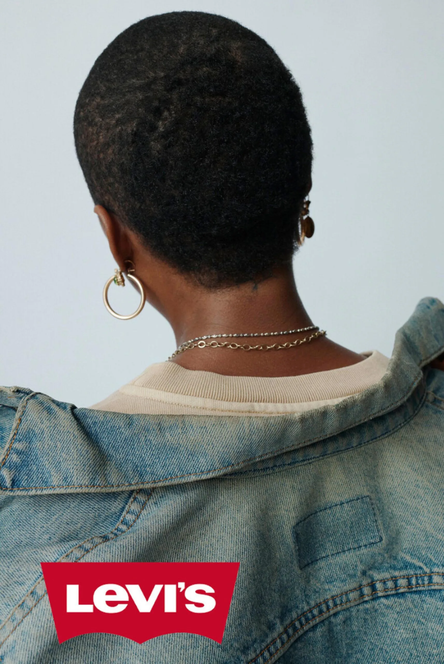 Model with cropped hair seen from behind, wearing a faded denim jacket, gold hoops and chain necklaces, for Levi's.