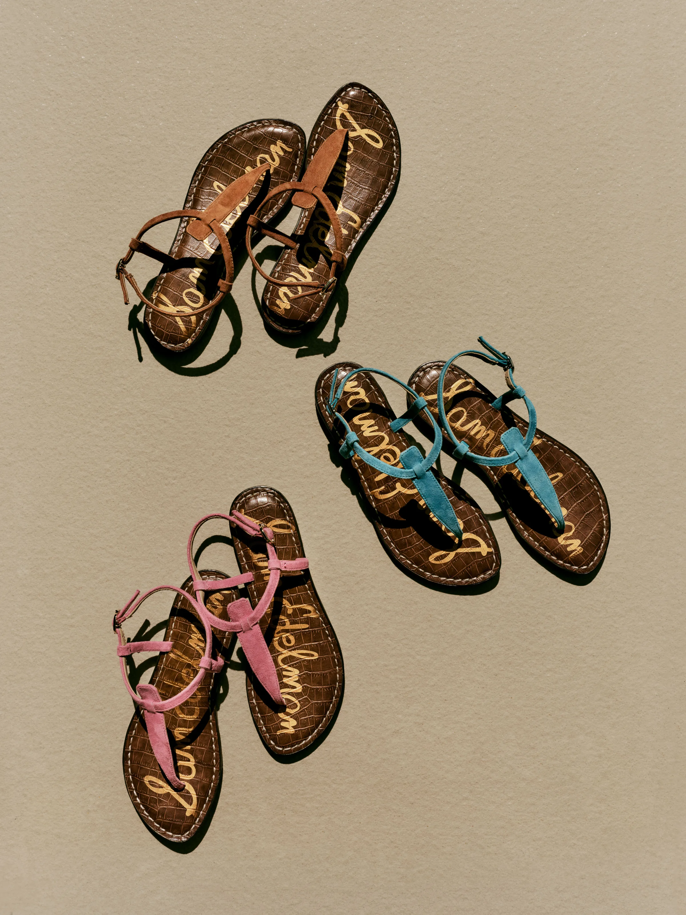 Three pairs of suede T-strap thong sandals in tan, teal and pink on a beige backdrop with hard shadows, for Sam Edelman.