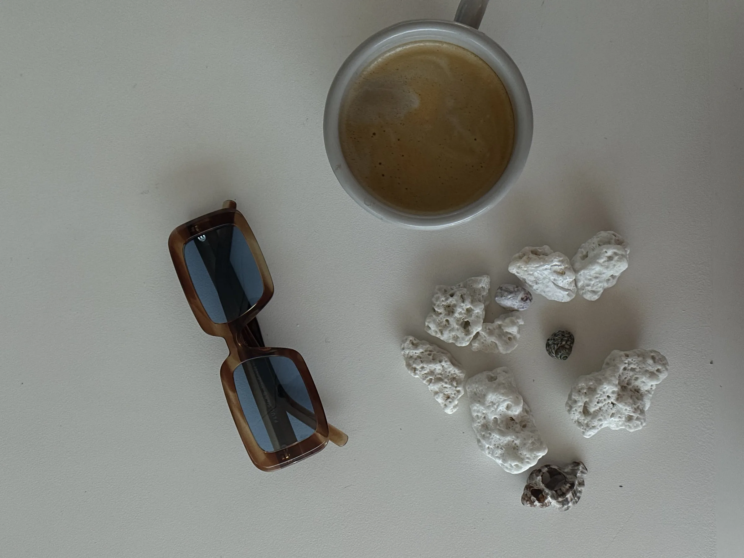 Tortoiseshell sunglasses with blue lenses beside a cup of coffee and white coral stones on a pale table.