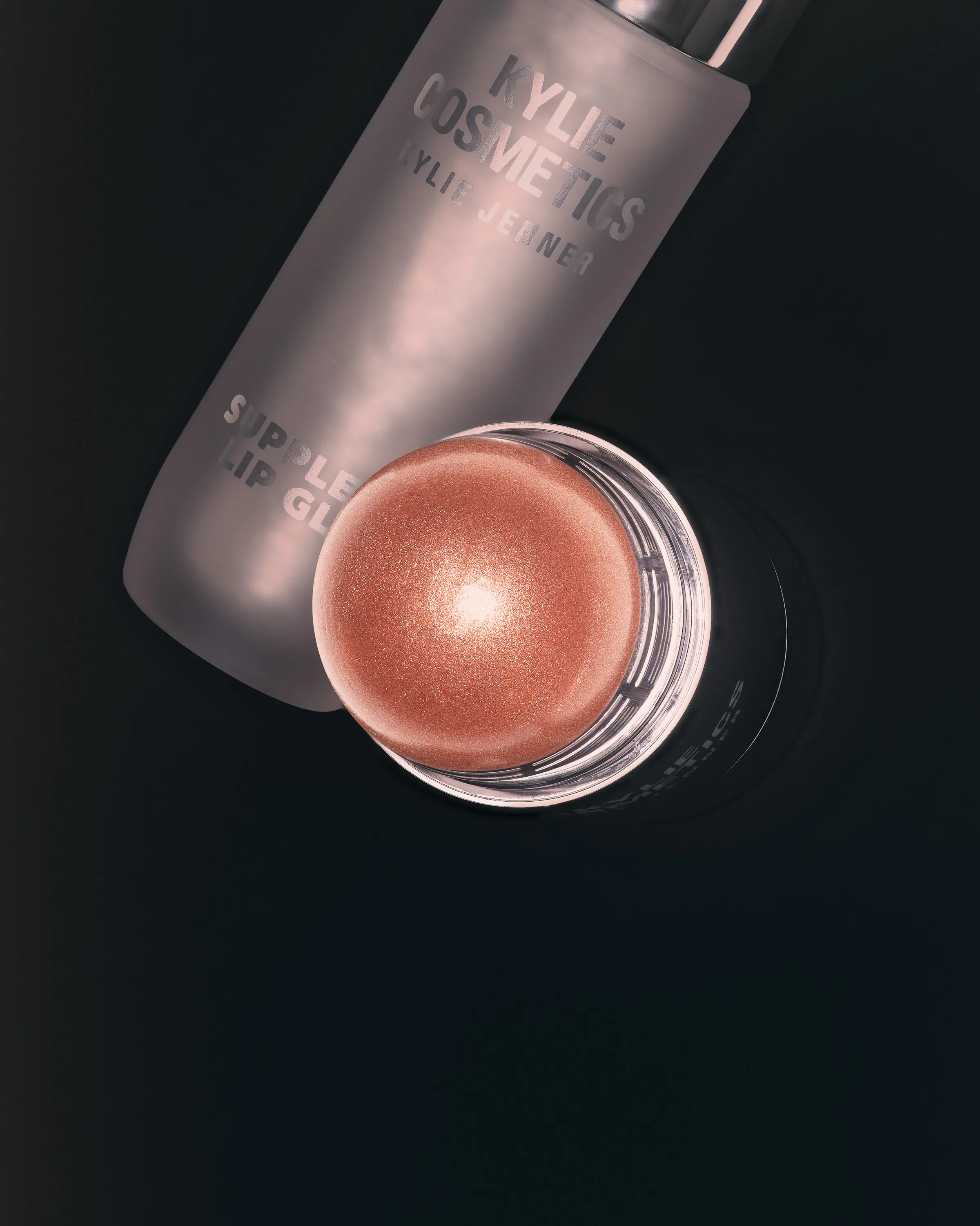 Frosted Supple Lip Glaze bottle and shimmering peach dewy balm stick on a black background, for Kylie Cosmetics.