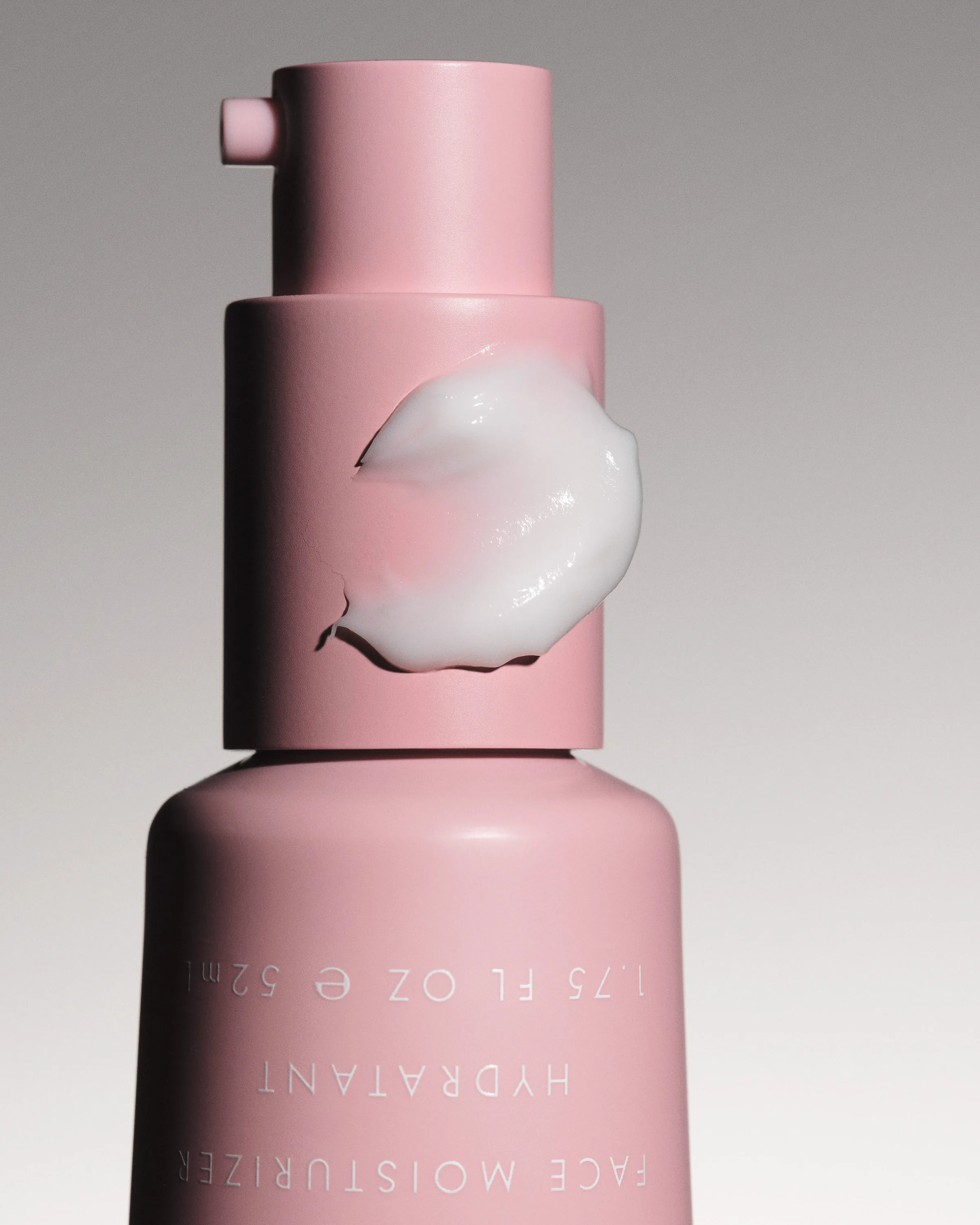 Pink face moisturizer pump bottle, upside down, with a white cream swatch on its neck against grey, for Kylie Cosmetics.