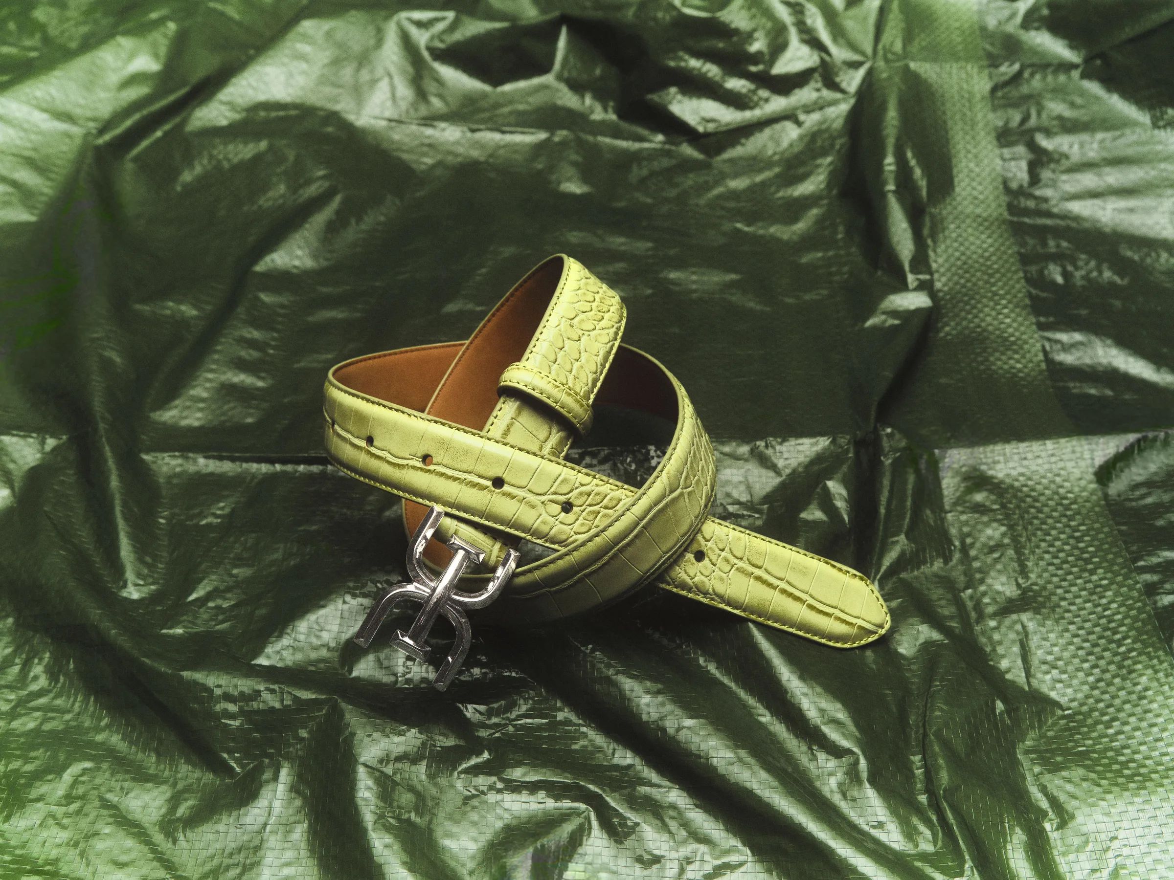 Light green croc-embossed leather belt with silver logo buckle coiled on crinkled green plastic, for Sam Edelman.