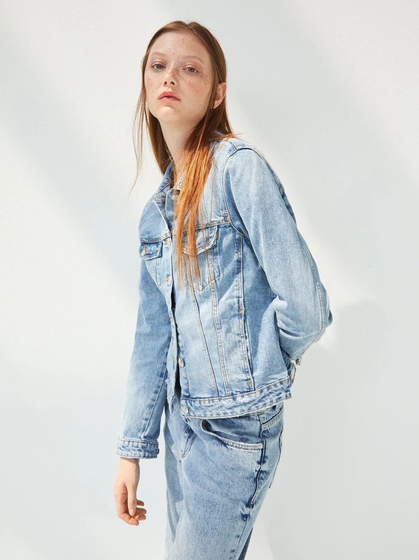 Red-haired model in a light wash denim jacket and matching jeans against a white wall with soft shadows, for Zara.