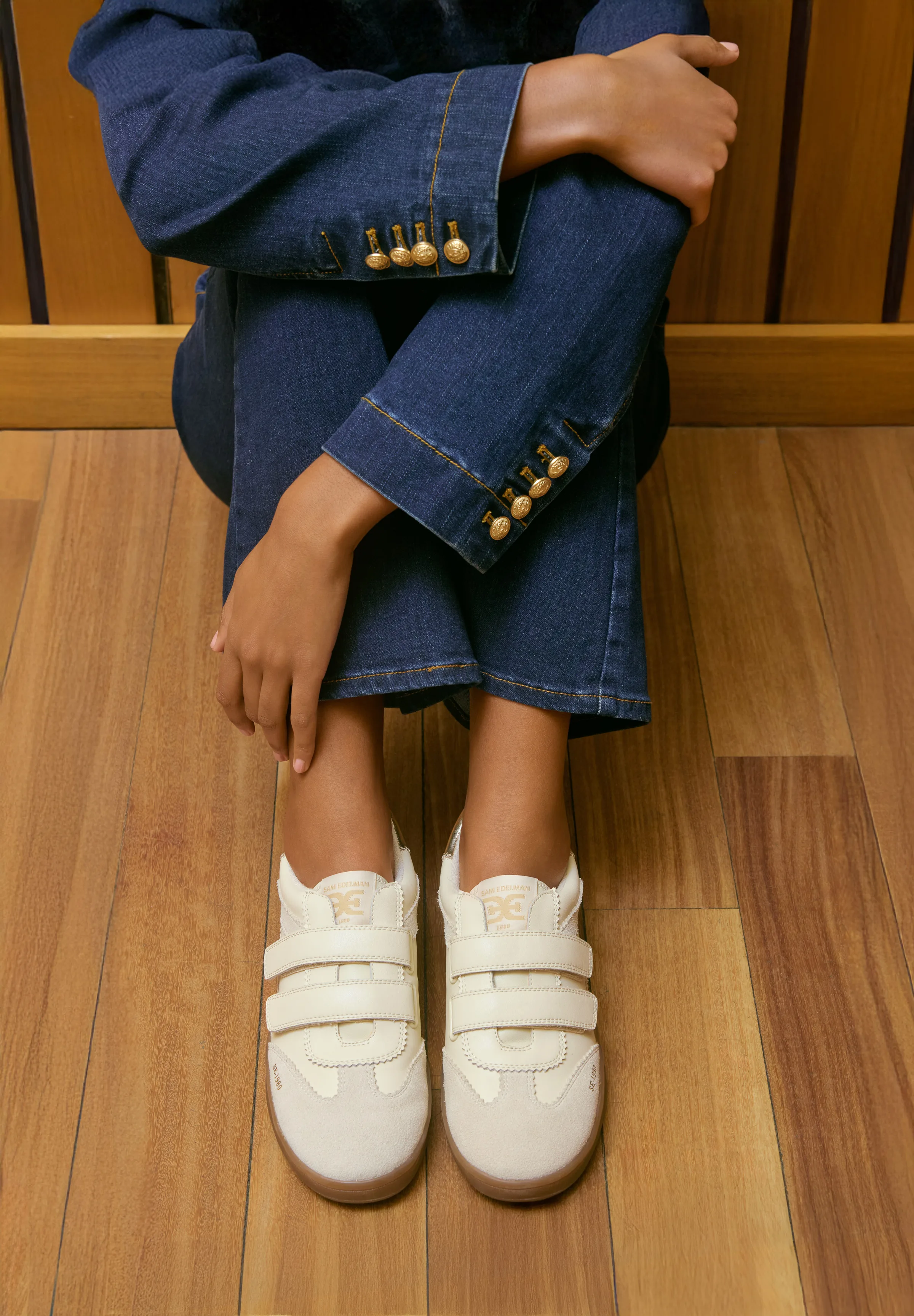 Seated model in a denim suit with gold buttons wearing ivory velcro-strap sneakers on a wooden floor, for Sam Edelman.