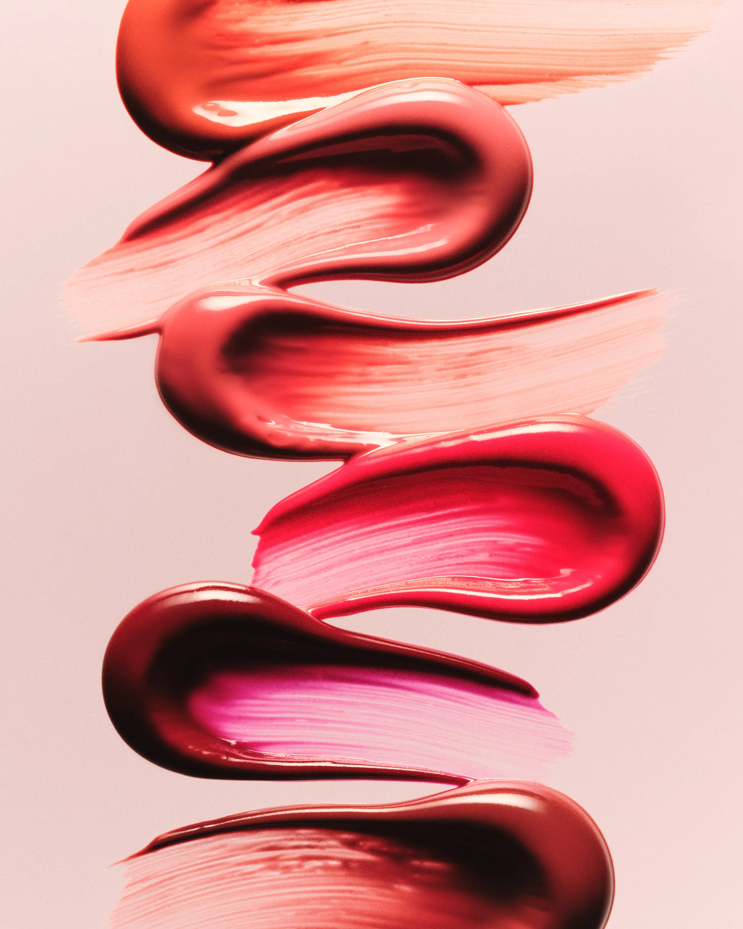 Stacked glossy lip product swatches in coral, red, pink and berry shades on a pale pink background, for Kylie Cosmetics.