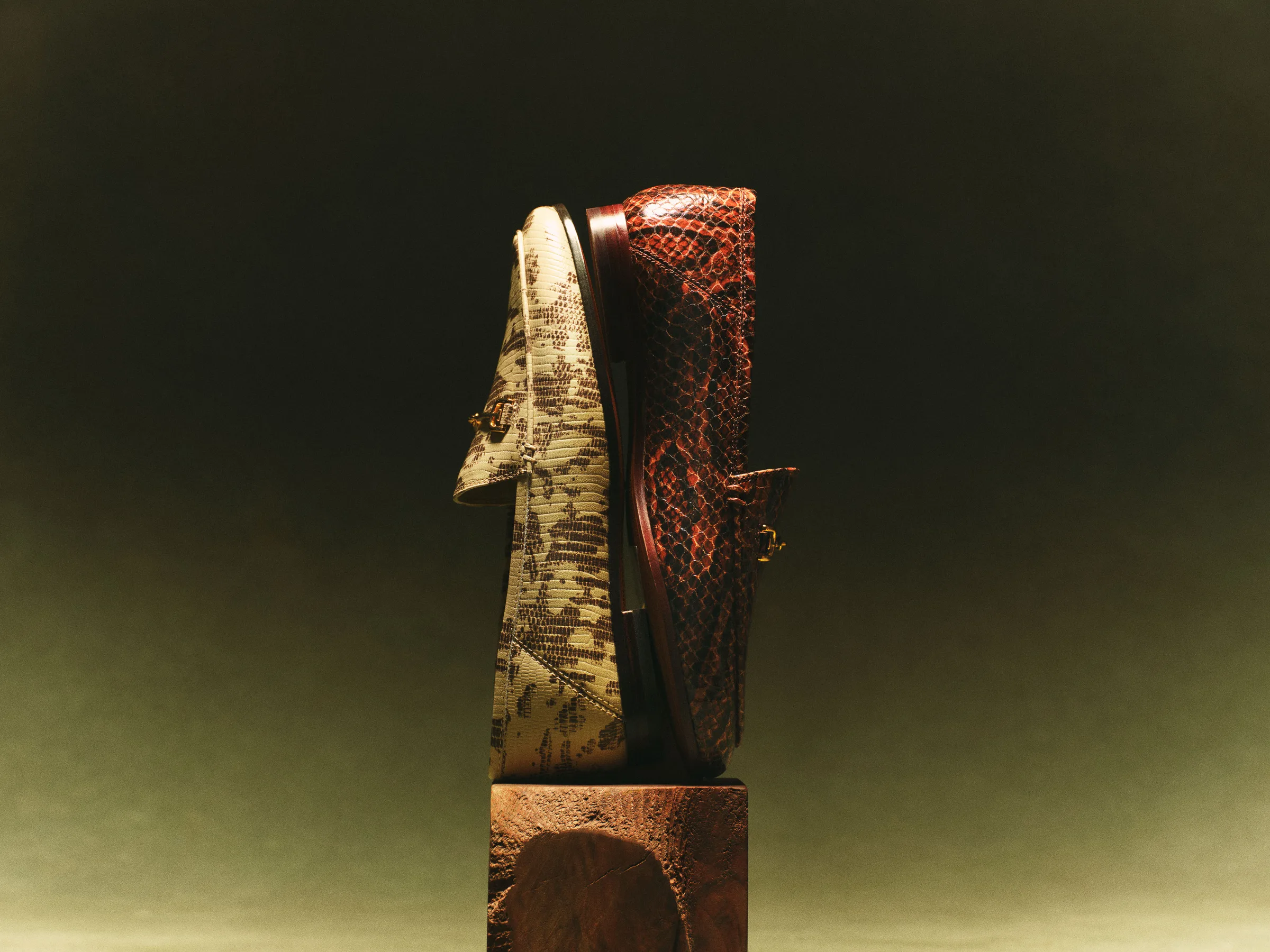 Two snakeskin-embossed loafers, beige and rust brown, stacked upright on a wooden block against dark green, for Sam Edelman.