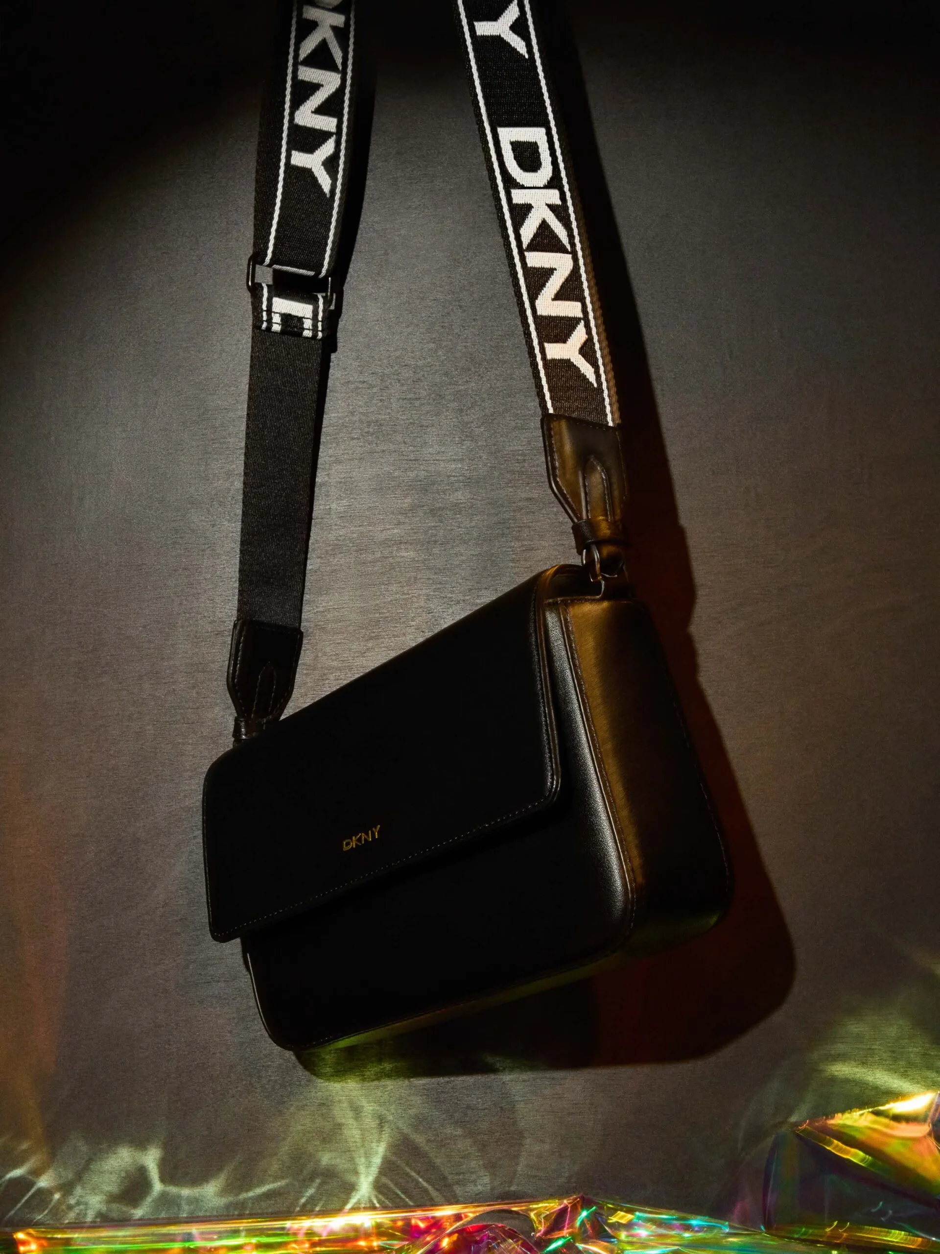 Black leather crossbody bag hanging from a logo webbing strap above iridescent film on a dark backdrop, for DKNY.