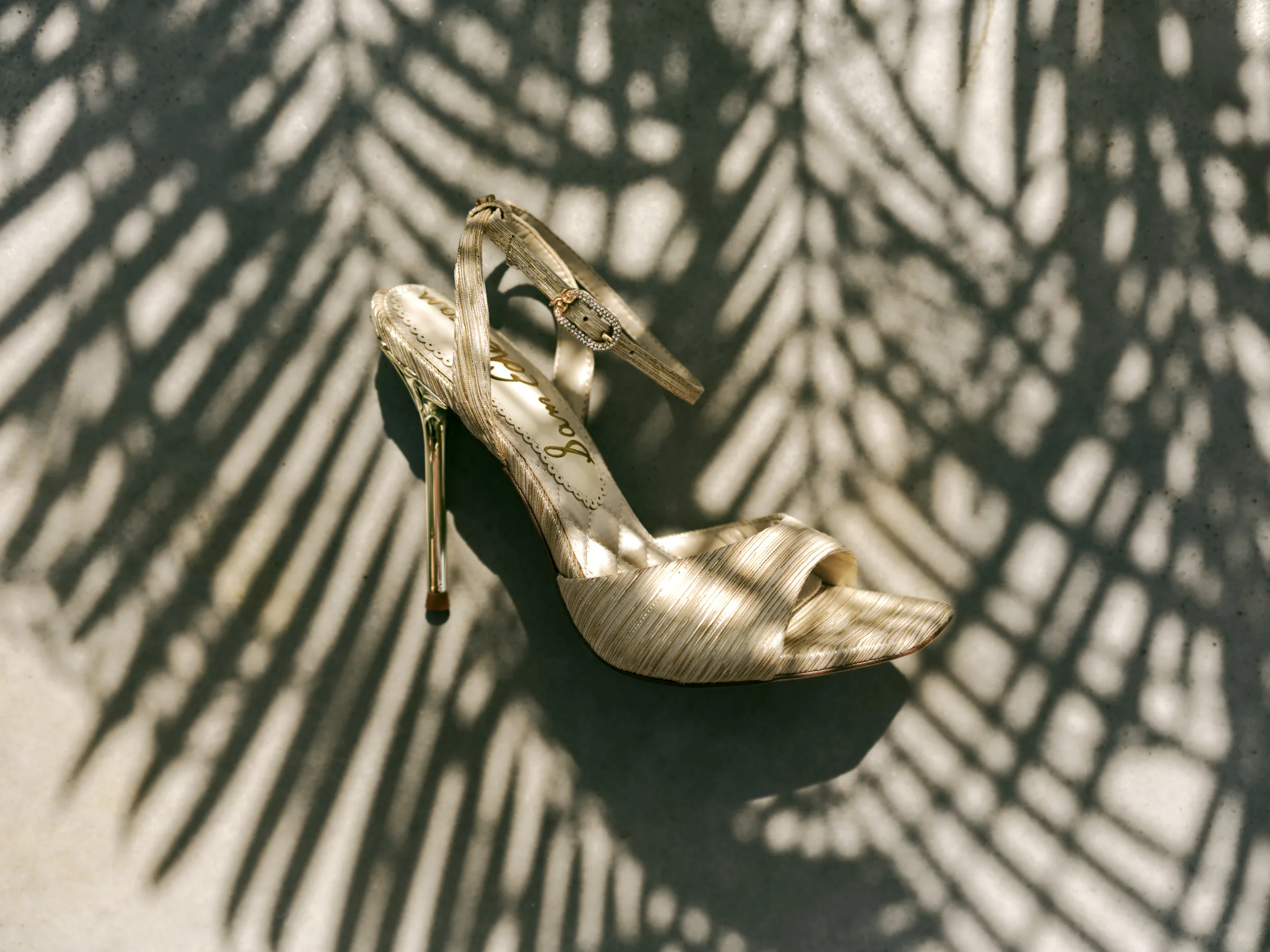 Gold pleated ankle-strap stiletto sandal on concrete under palm leaf shadows, for Sam Edelman.