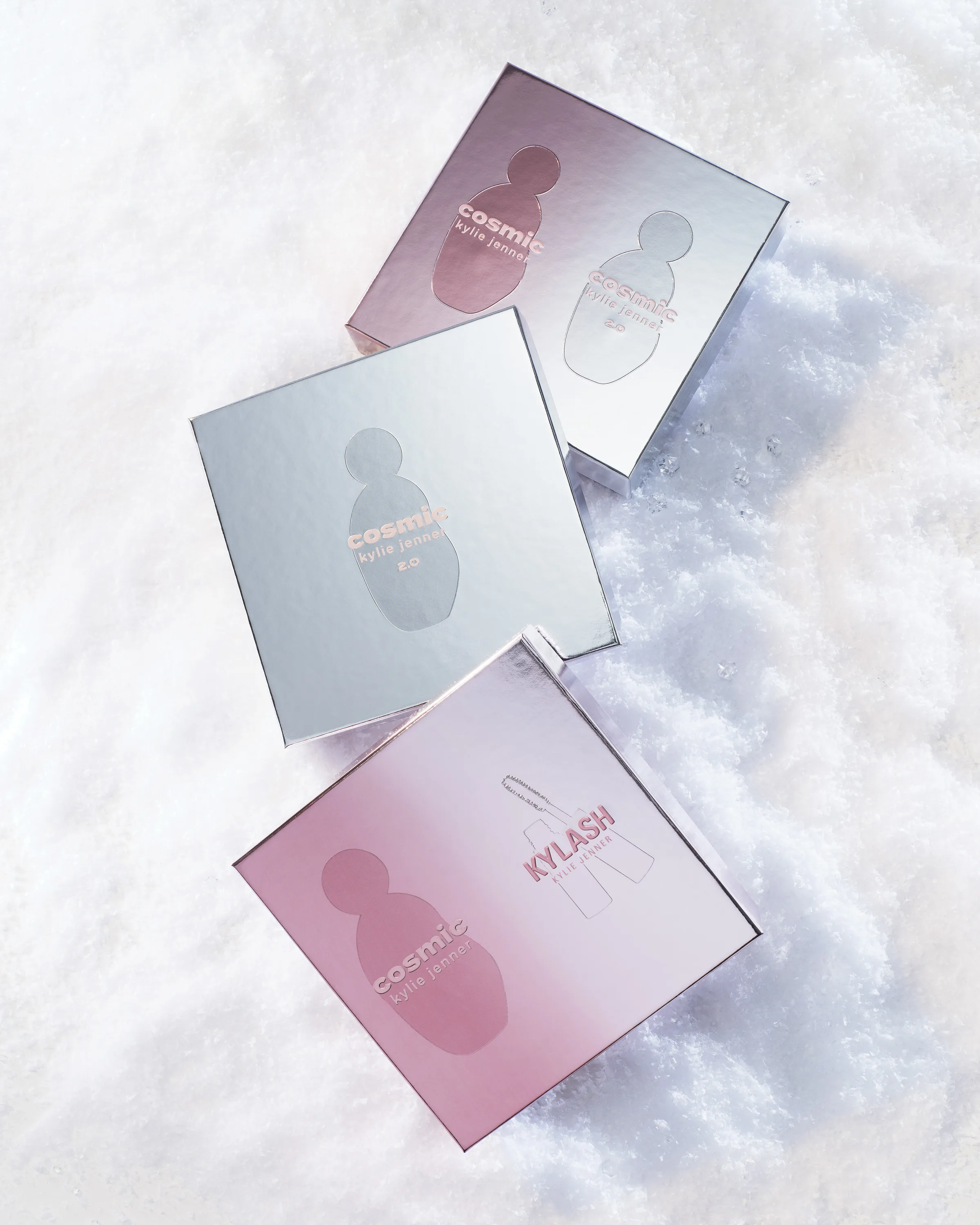 Three metallic pink and silver Cosmic Kylie Jenner fragrance gift boxes resting in snow, for Kylie Cosmetics.