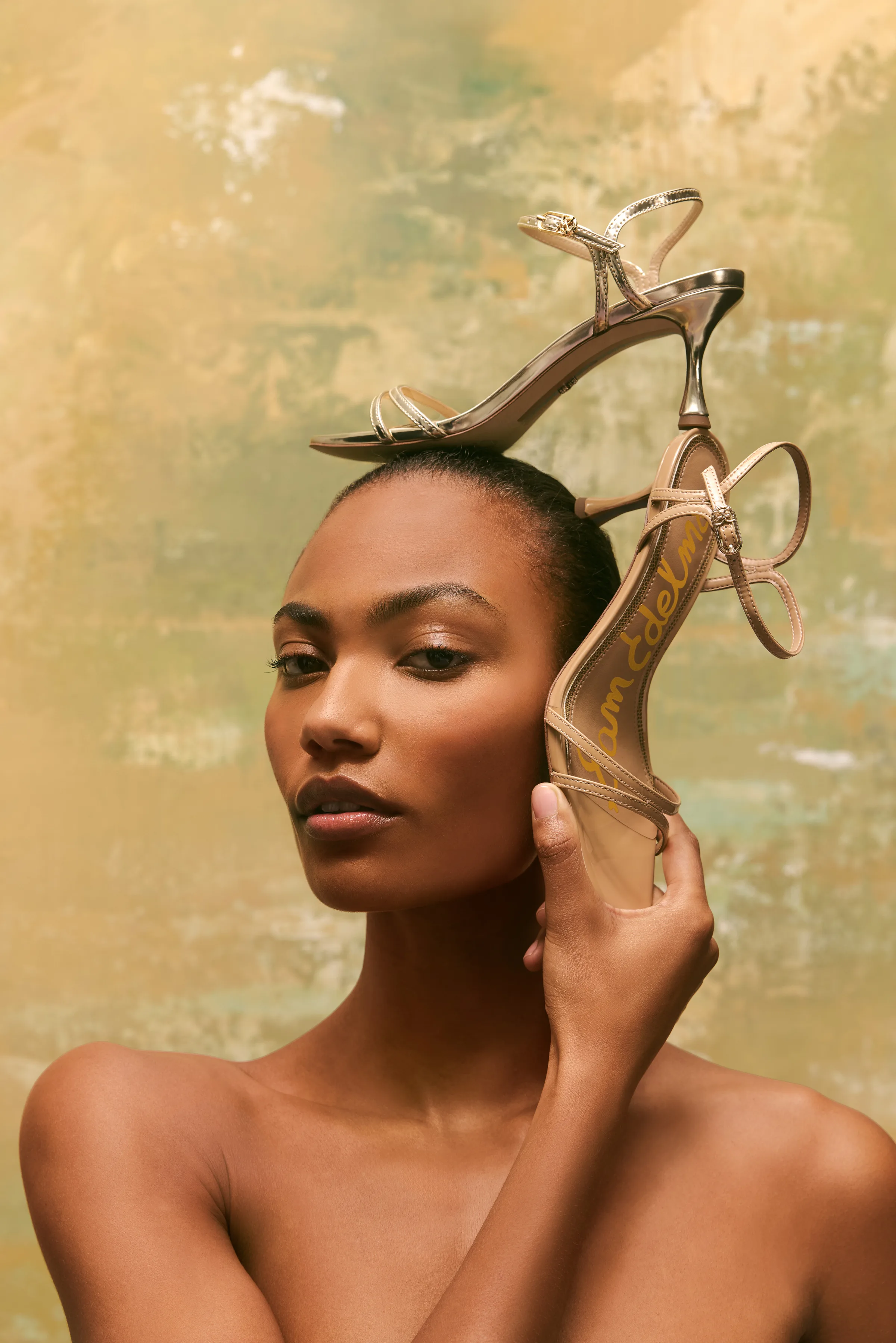 Model balancing gold strappy heeled sandals on her head against a painted gold-green backdrop, for Sam Edelman.