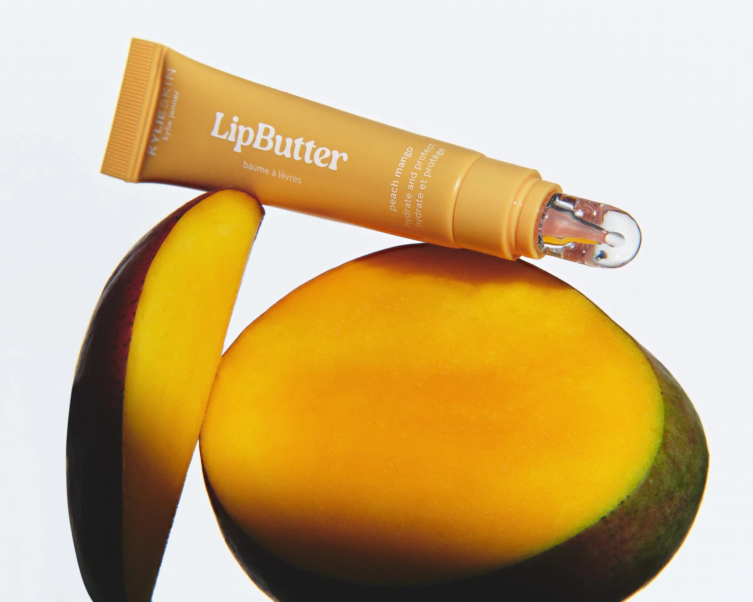Peach mango Kylie Skin LipButter tube balanced on halved mango slices against a white background, for Kylie Cosmetics.