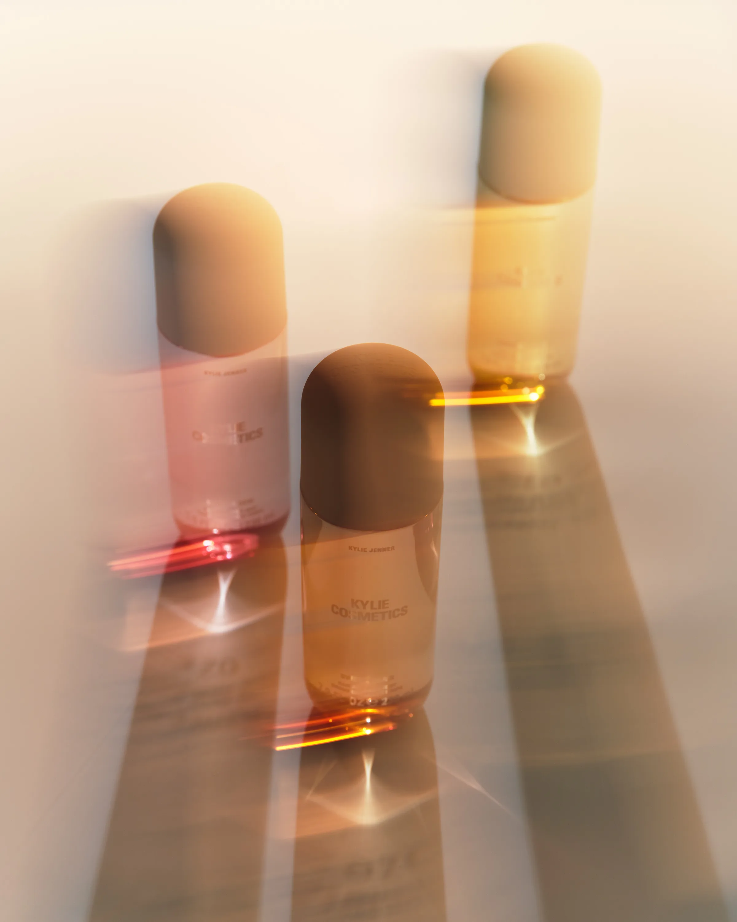Three body mist bottles with rounded caps casting long shadows and golden light refractions on a white surface, for Kylie Cosmetics.