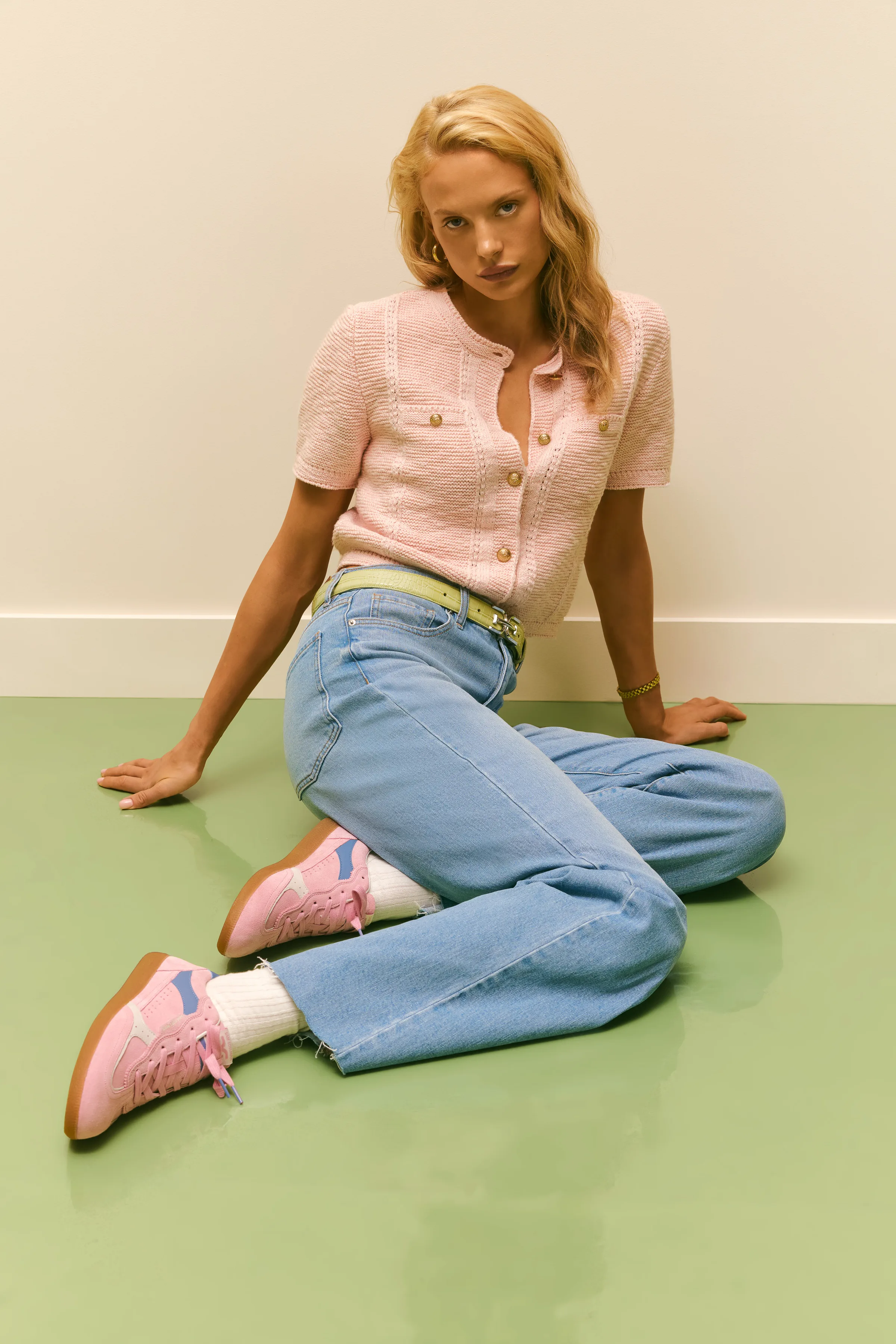 Model seated on a green floor in a pink knit top, light jeans and pink suede sneakers, for Sam Edelman.