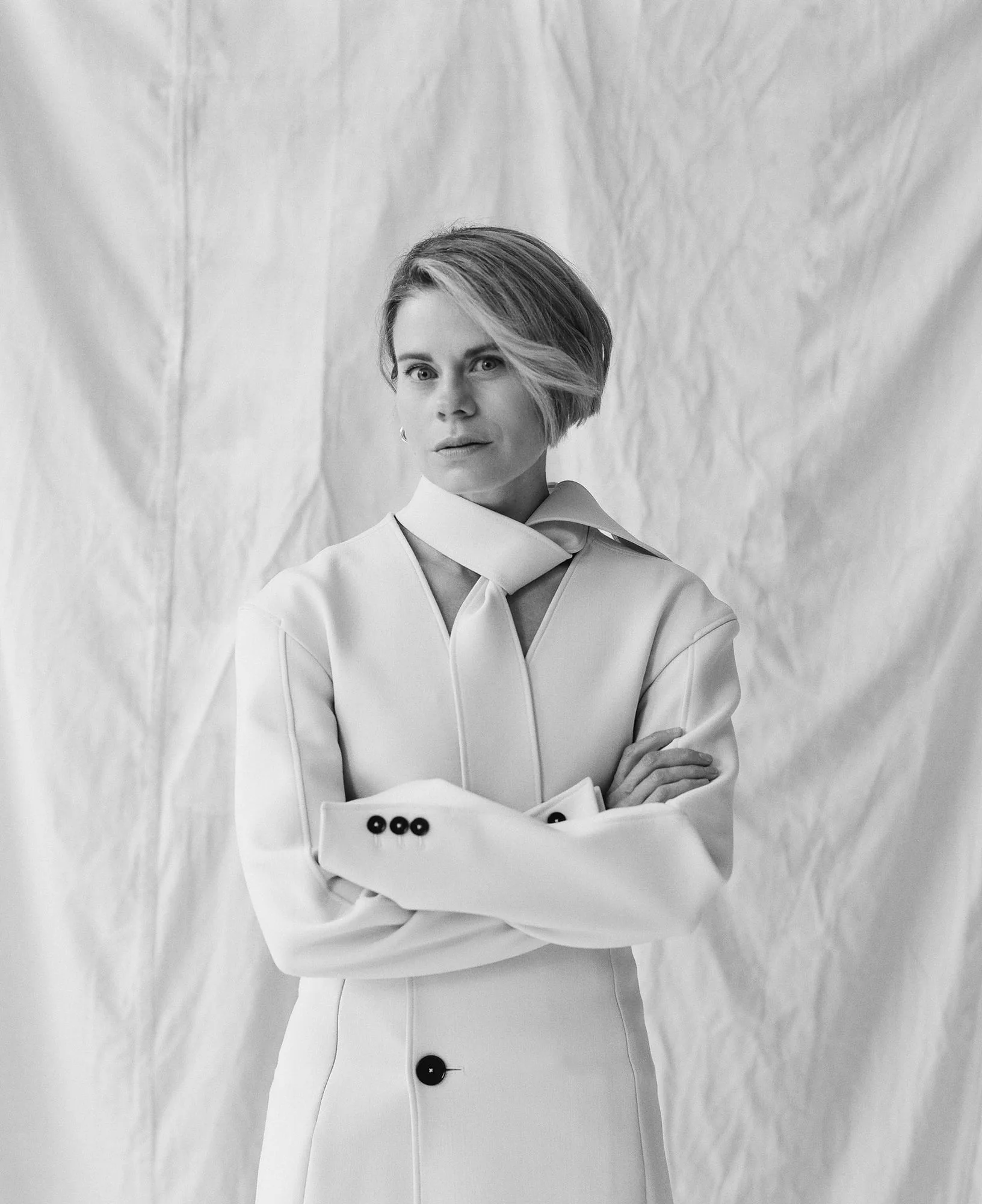 Black and white portrait of a woman in a white coat with tie collar, arms crossed before a creased backdrop, for The Last Magazine.