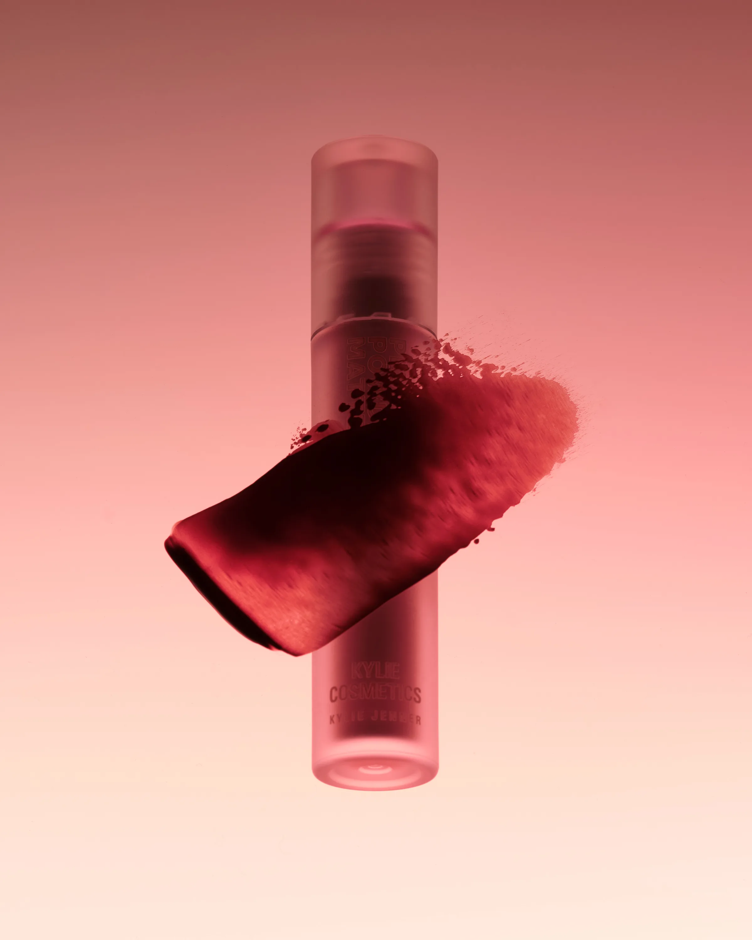 Frosted lipstick tube with a red smeared swatch across it on a pink gradient background, for Kylie Cosmetics.