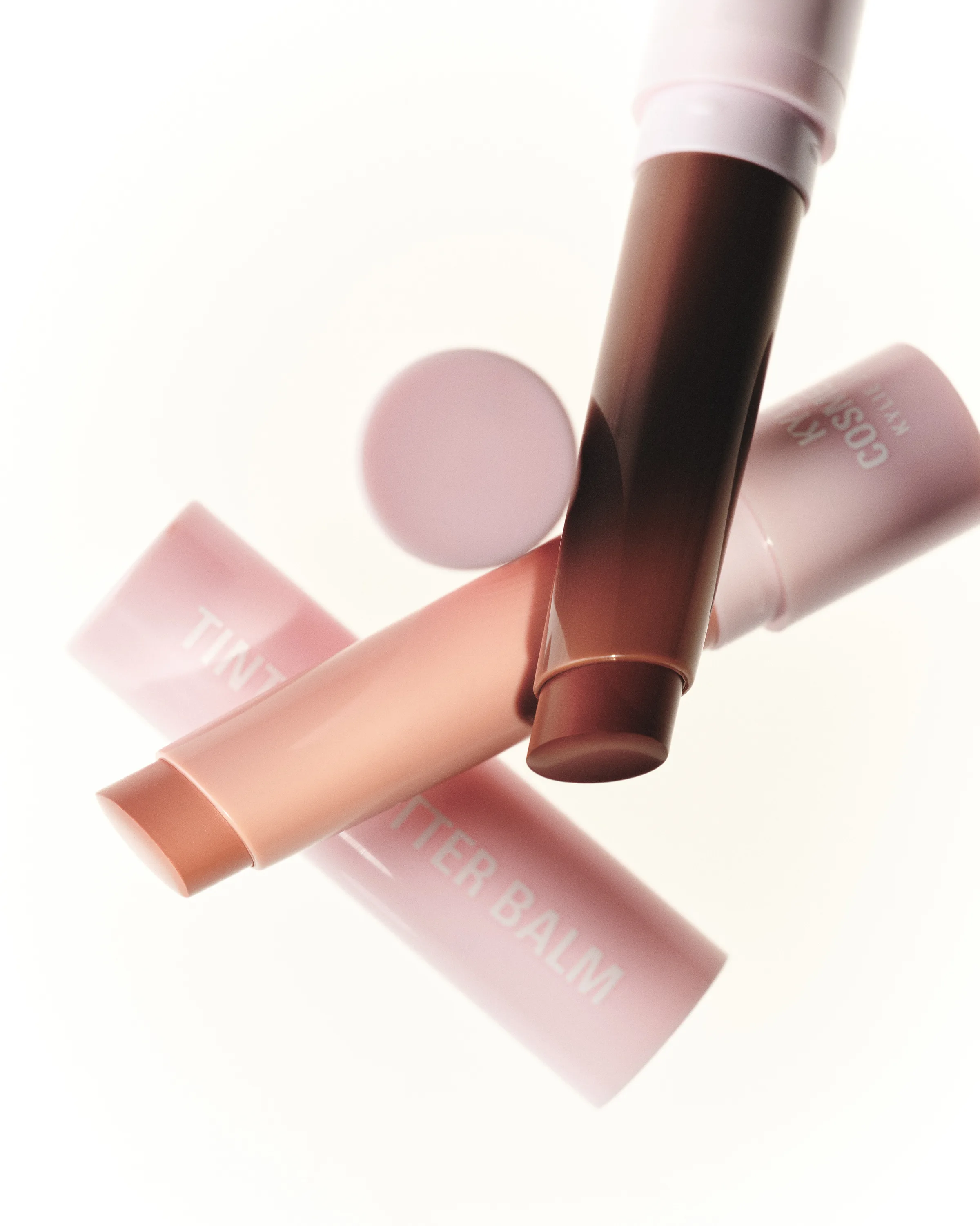 Still life of Tinted Butter Balm sticks in Mocha Kiss and Angel Cake with pink tubes on white, for Kylie Cosmetics.