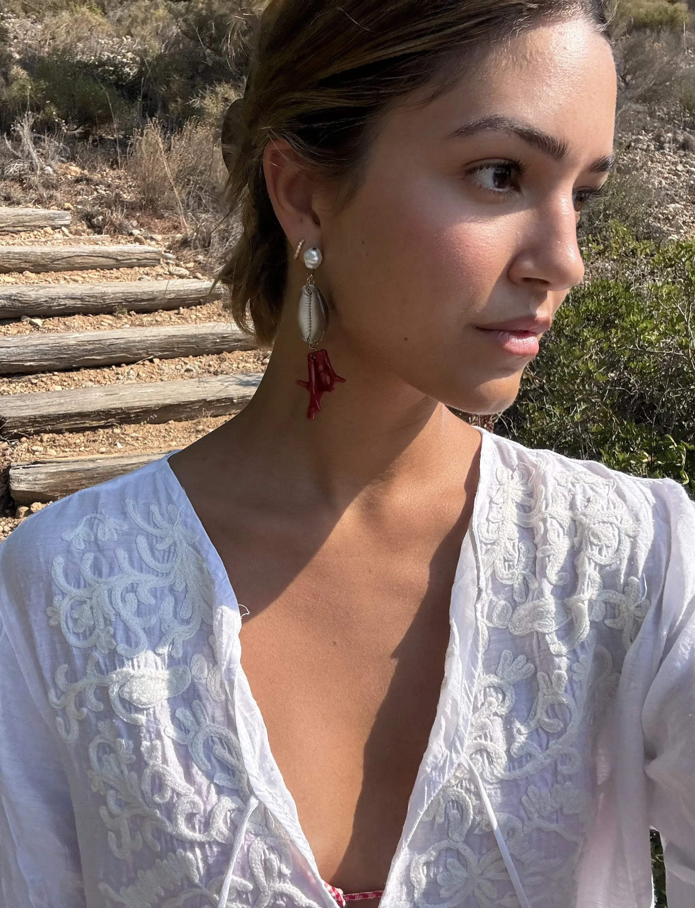 Woman in a white embroidered tunic wearing pearl, cowrie shell and red coral drop earrings on a sunny hillside path.