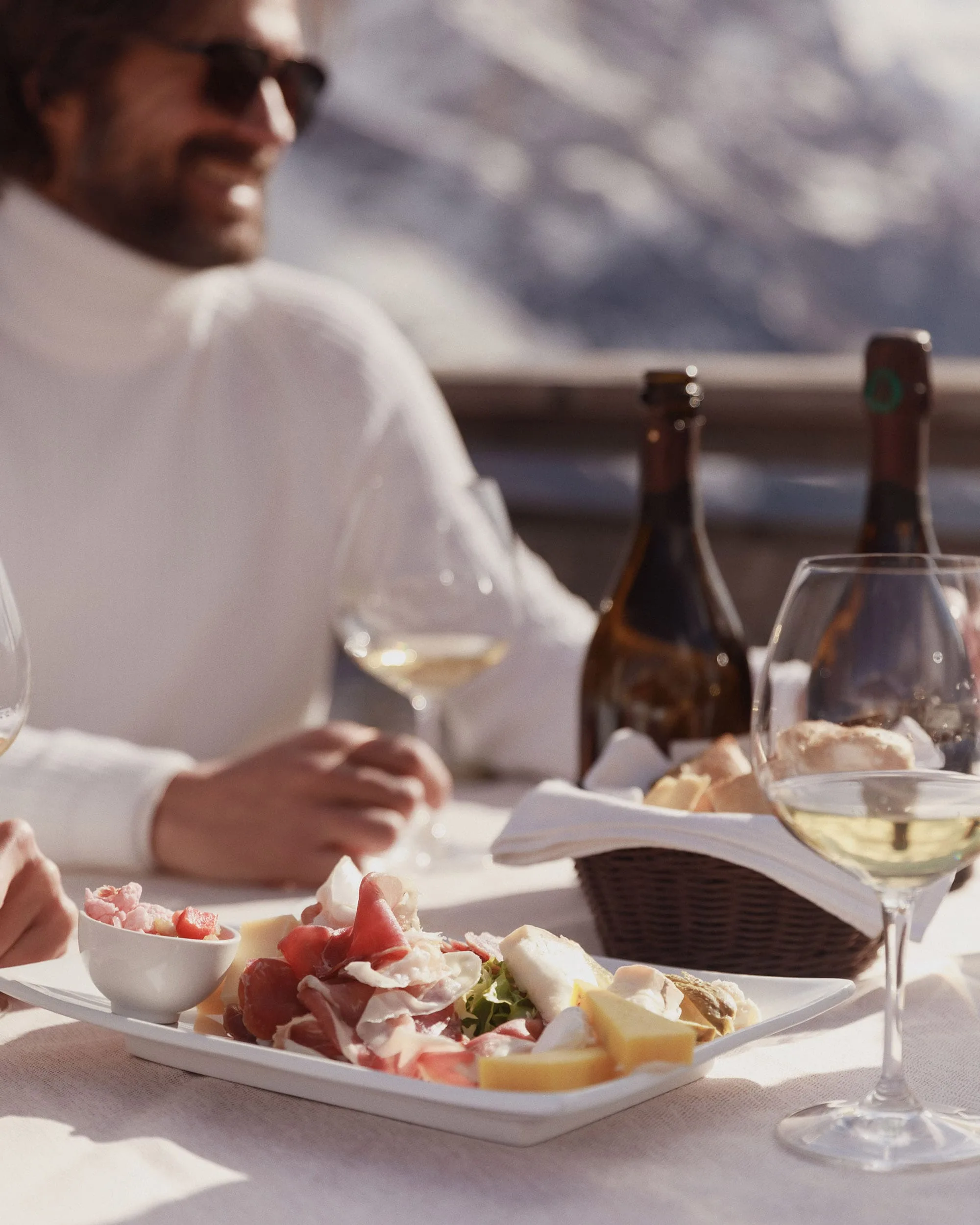 Man in white turtleneck at a sunny mountain terrace table with charcuterie, cheese, bread and white wine, for Marriott.