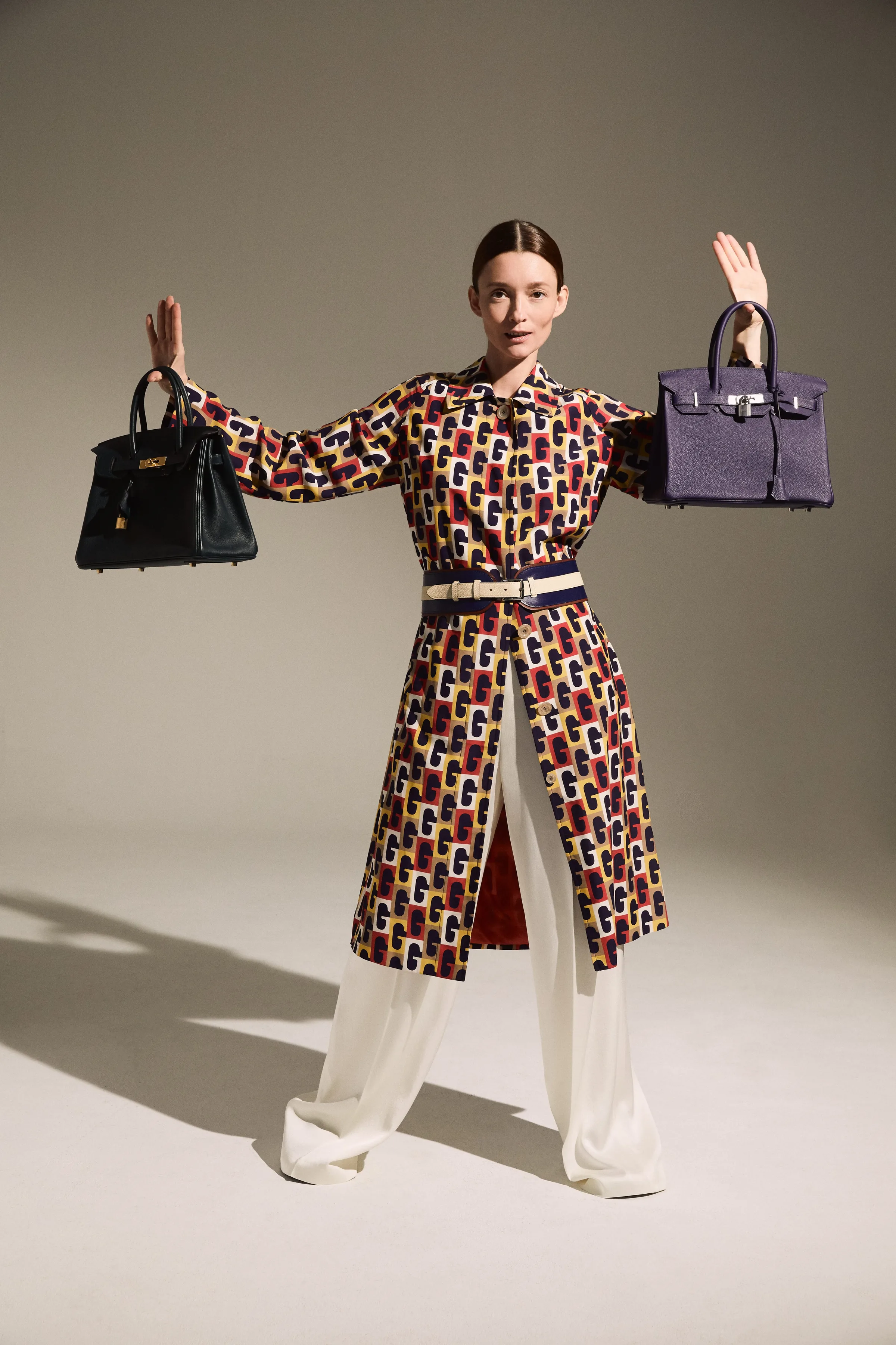 Model in a printed coat and white trousers holds a black and a purple leather handbag aloft, for The RealReal.