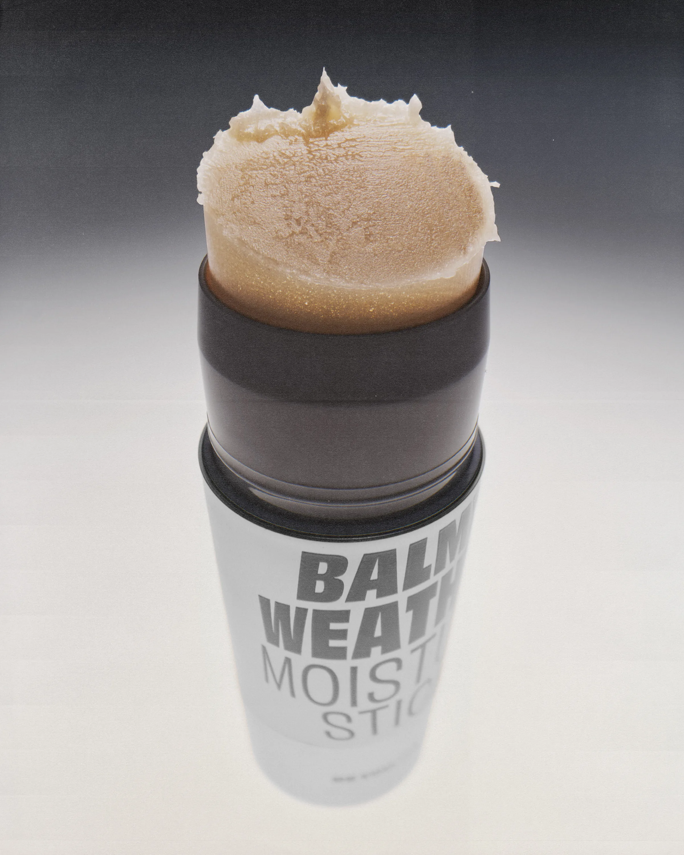 Open Balm Weather moisture stick with shimmering golden balm on a grey gradient backdrop, for Good Weird.