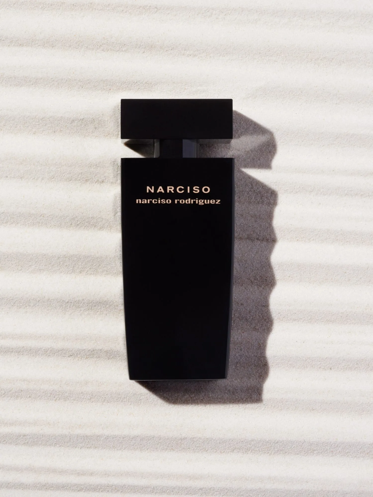 Black Narciso perfume bottle lying on rippled white sand with long shadows, for Narciso Rodriguez.