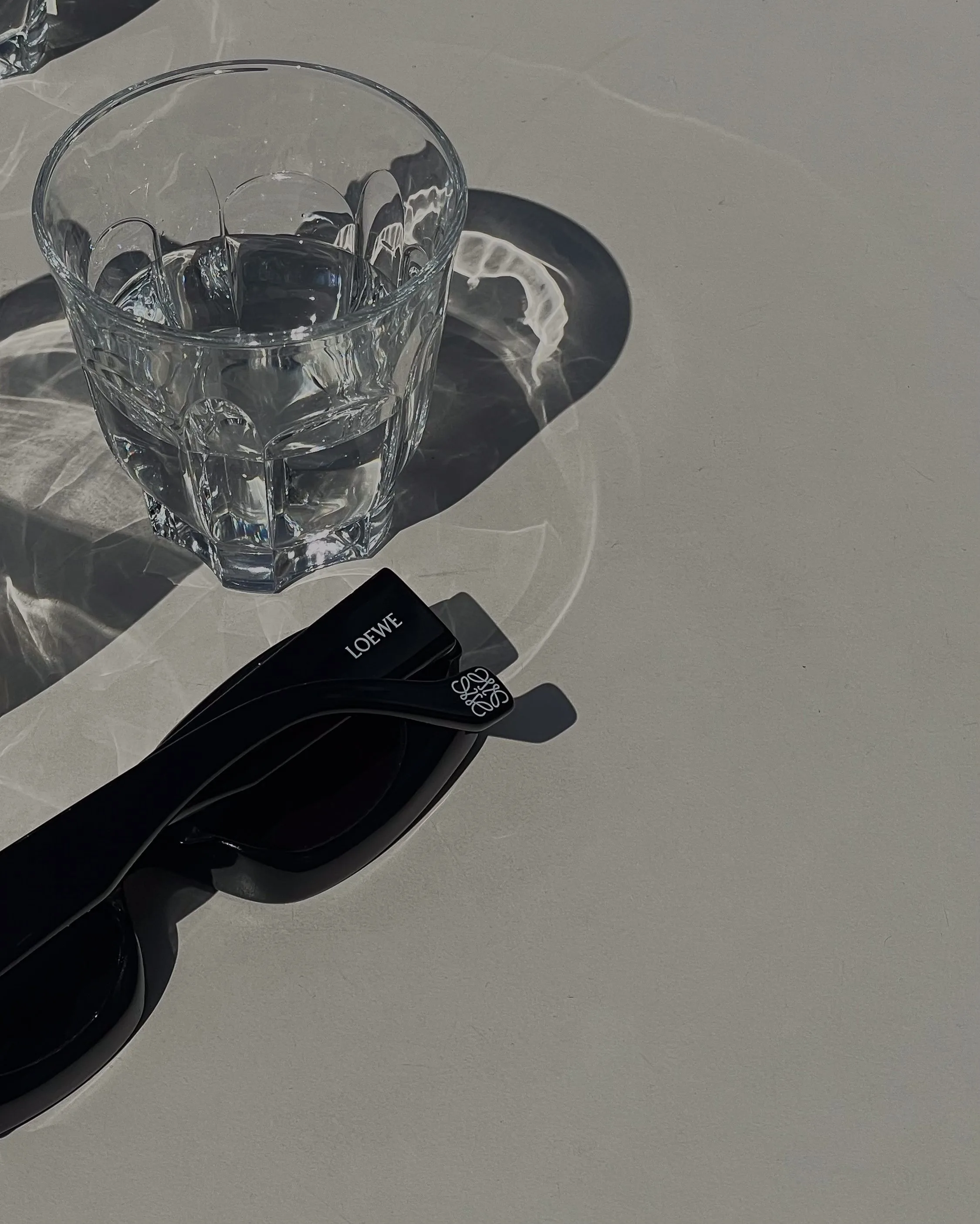 Black sunglasses with anagram temple beside a cut-glass tumbler of water in harsh sunlight on a white table, for Loewe.