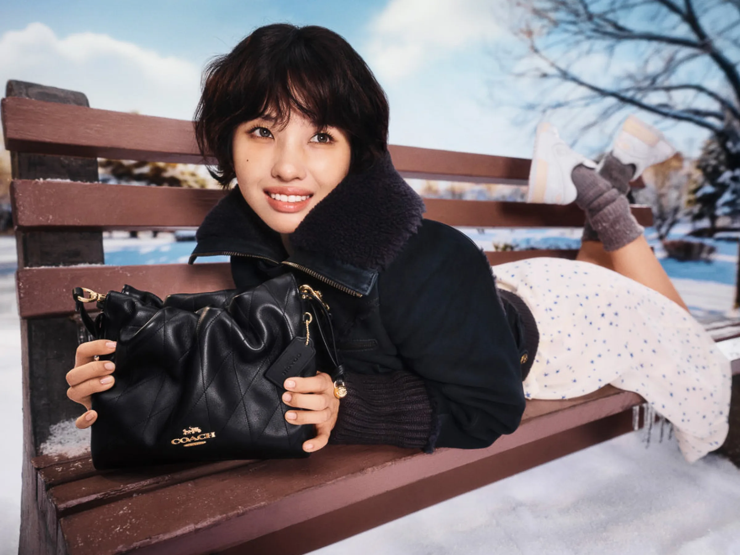 AI image of a woman lying on a snowy park bench holding a black quilted leather handbag, in a dark shearling jacket, for Coach.