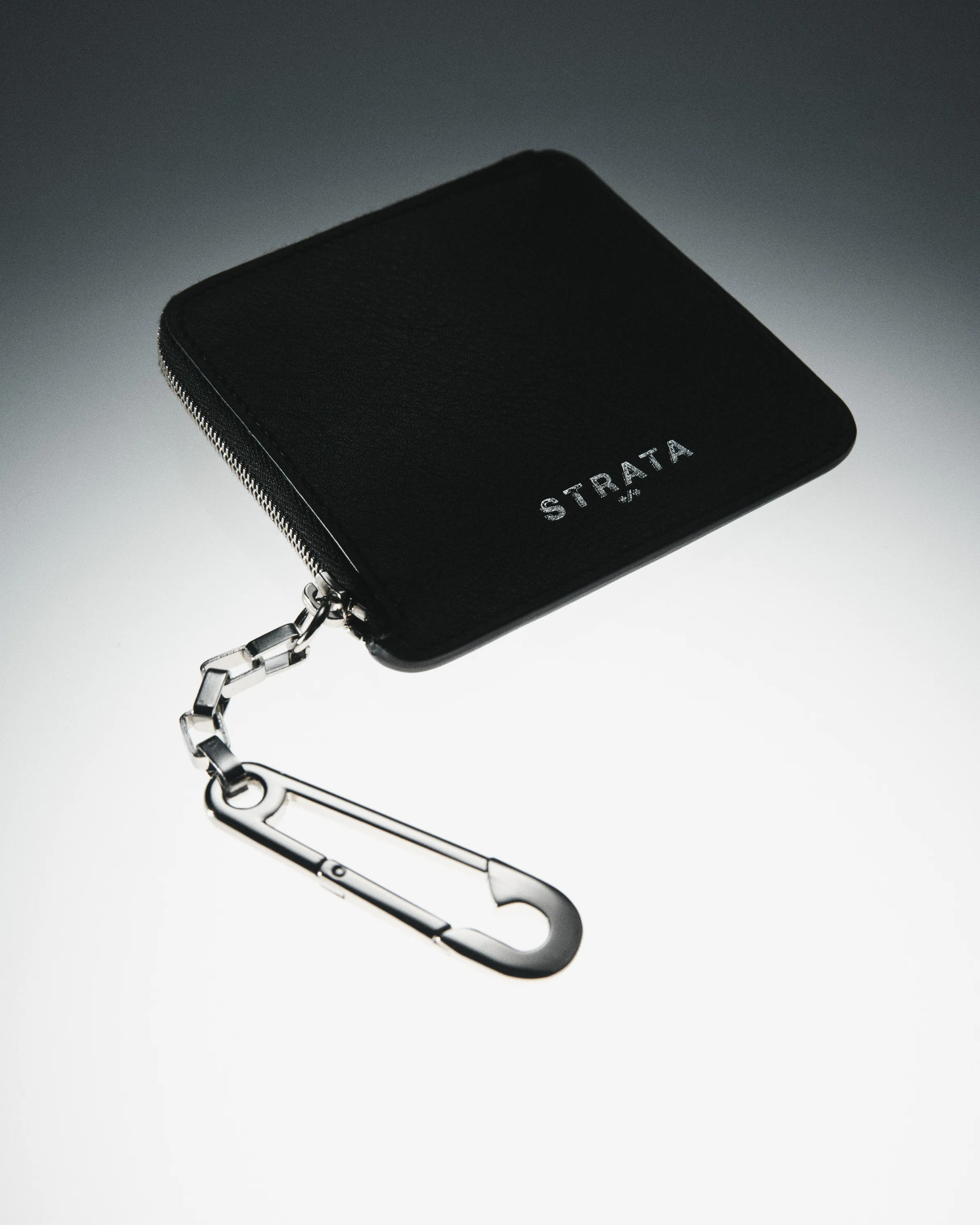 Black leather zip wallet with a silver chain and safety-pin clasp on a grey gradient backdrop, for Strata.
